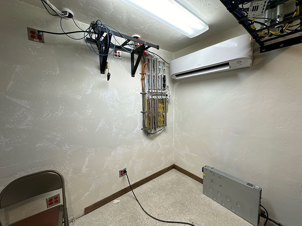 Small server room with a wall-mounted AC unit, cable rack, wiring, and a portable equipment case on the floor.