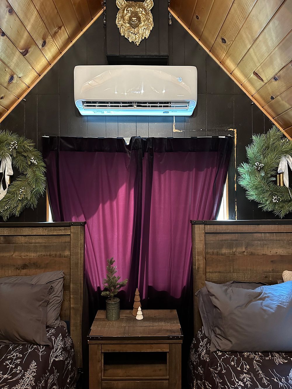 Two beds with gray bedding and wreaths on the wall, separated by a nightstand beneath a purple curtain and AC unit.
