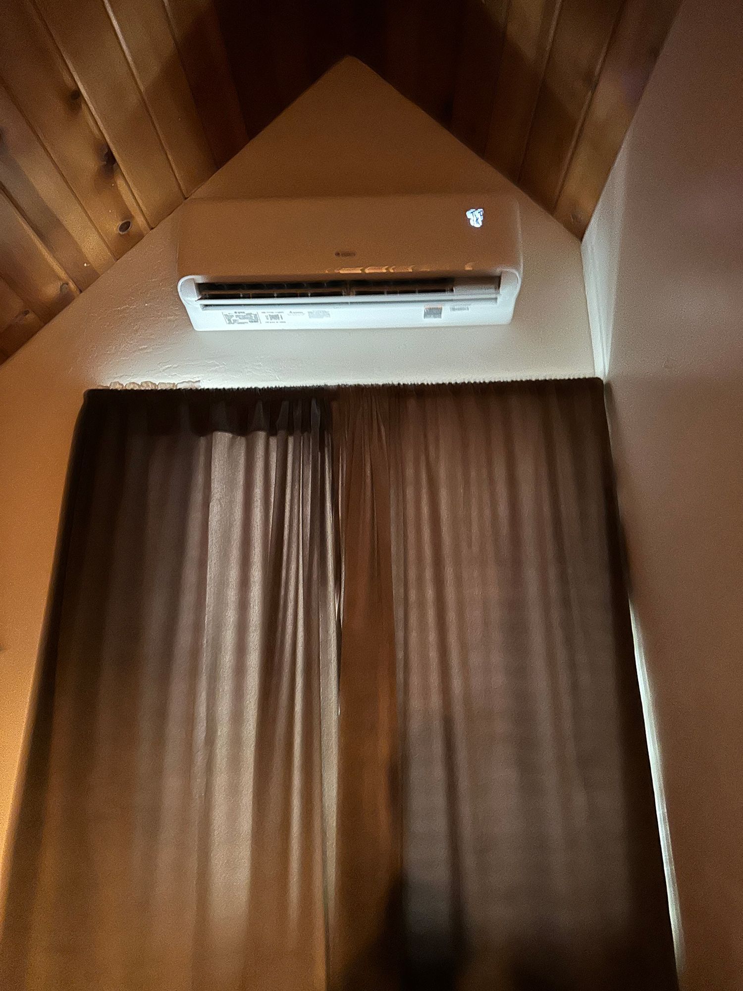 A white split-system air conditioner mounted on a wall above brown curtains under a wooden vaulted ceiling.