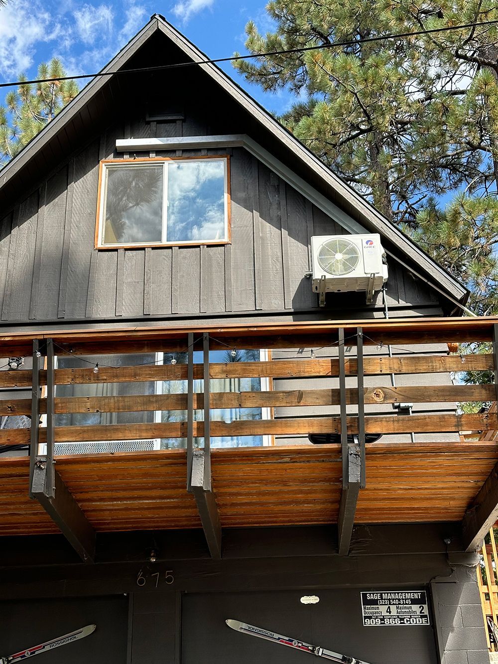 A dark-toned, A-frame cabin featuring a wooden balcony, an upstairs window, and an exterior air conditioning unit.
