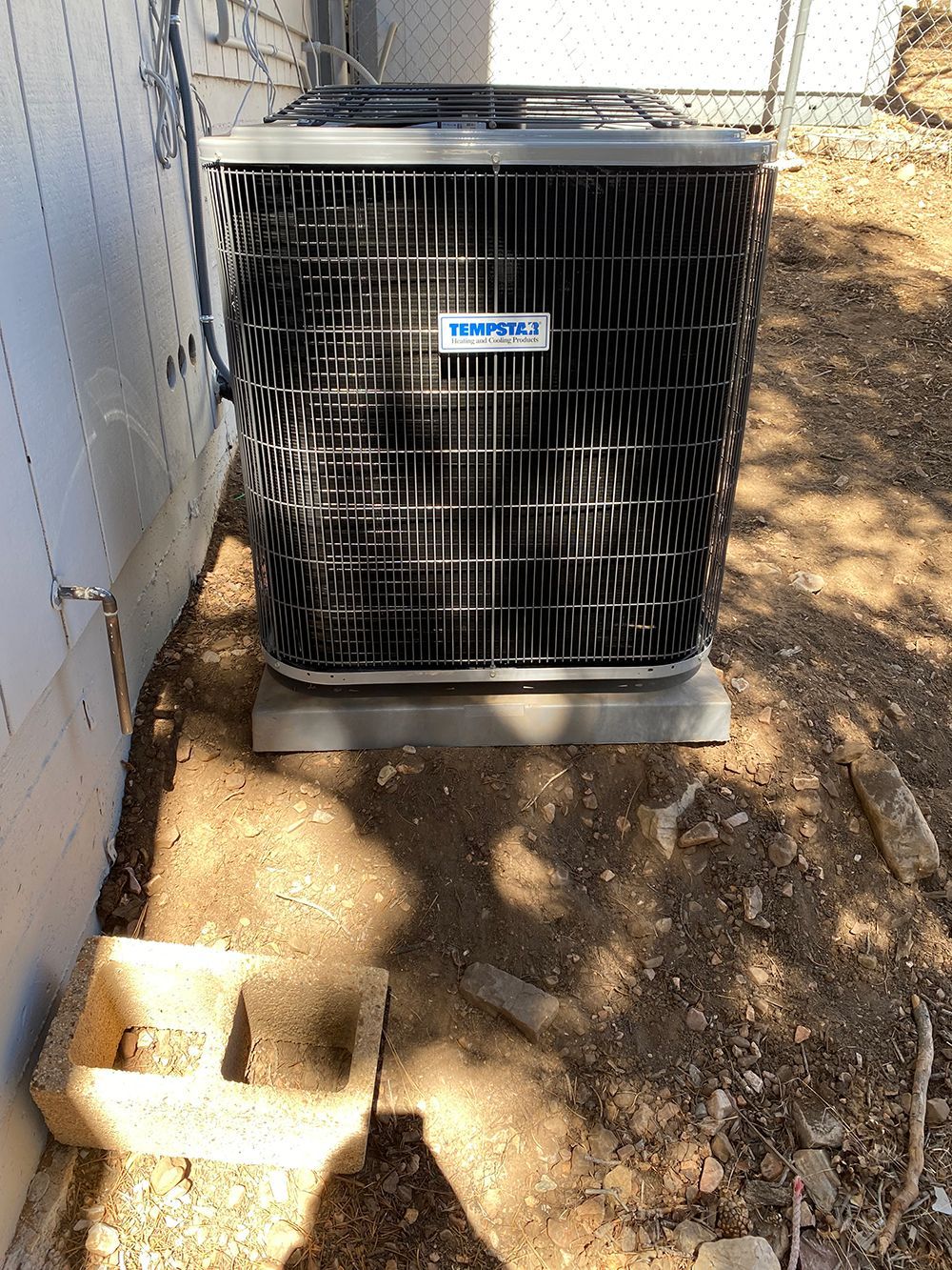 A Trane outdoor air conditioning condenser unit sits on a grey pad on dirt ground next to a white siding wall.