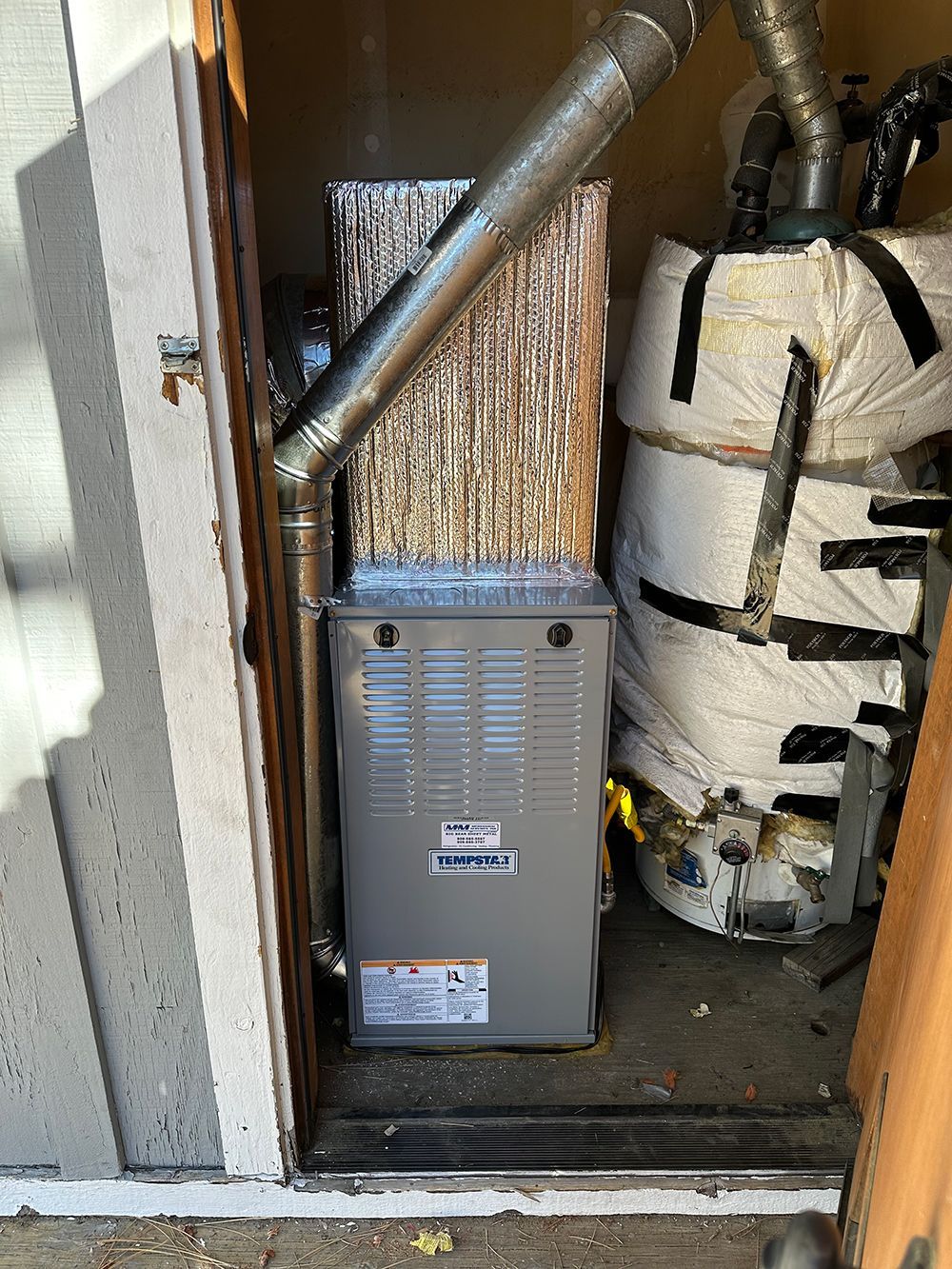 A gray HVAC furnace unit sits next to a water heater wrapped in white insulation inside a small utility closet.