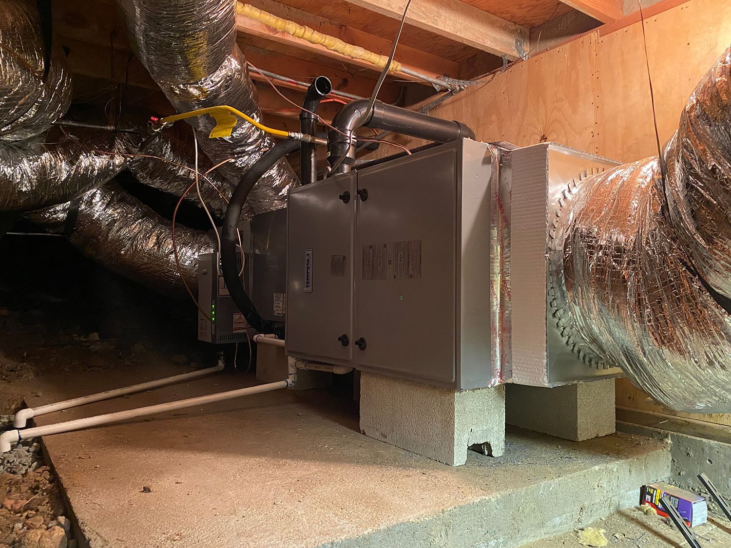 HVAC air handler unit installed on concrete blocks in a crawl space with attached silver insulated flexible ductwork.