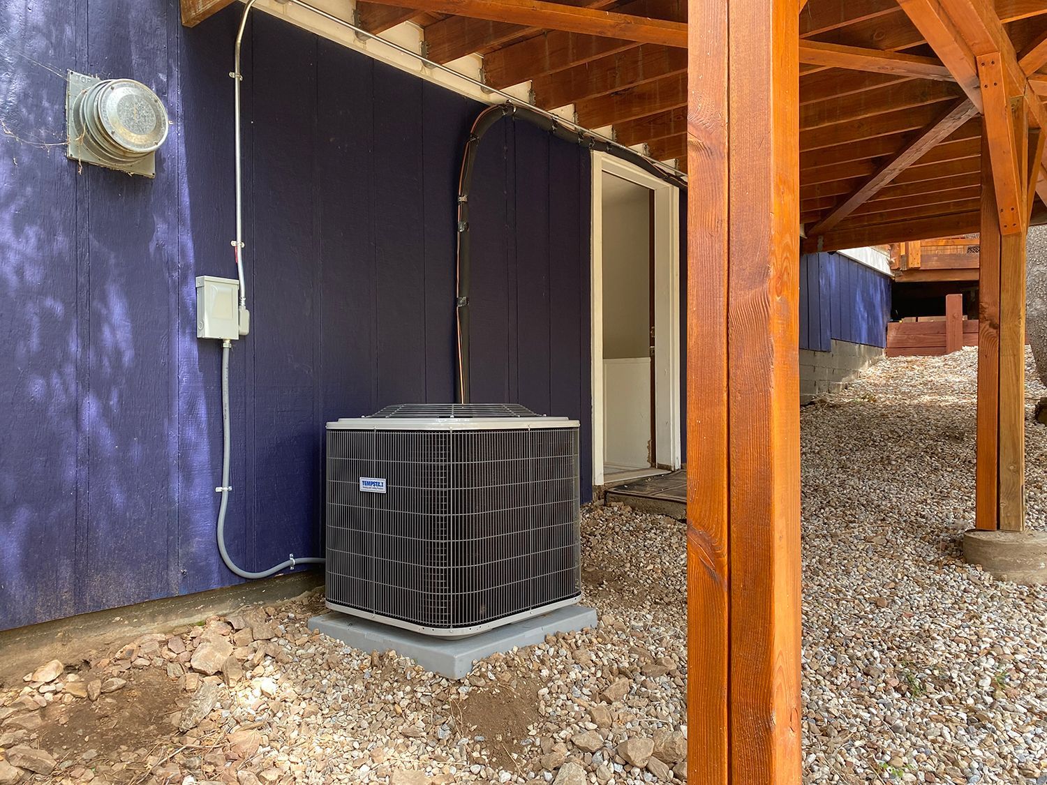 An HVAC outdoor unit sits on a gravel patch against a dark blue exterior wall, partially sheltered by a wooden deck.