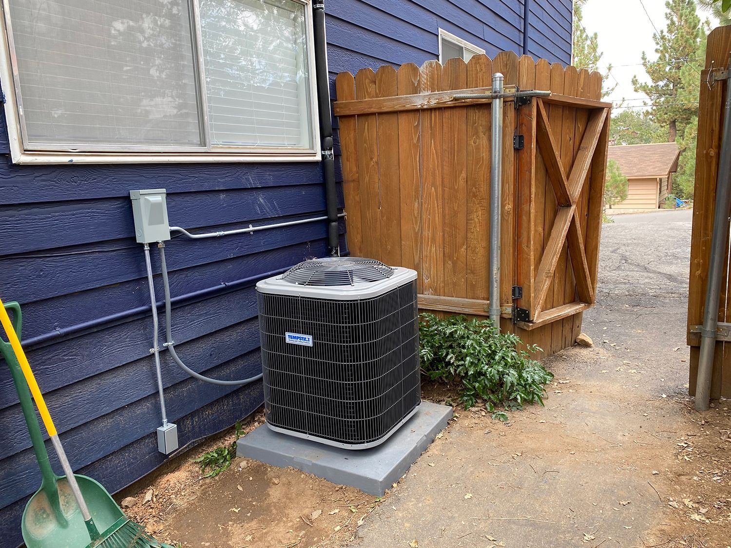 An HVAC outdoor unit on a concrete pad against a dark blue siding wall, next to a wooden privacy fence.