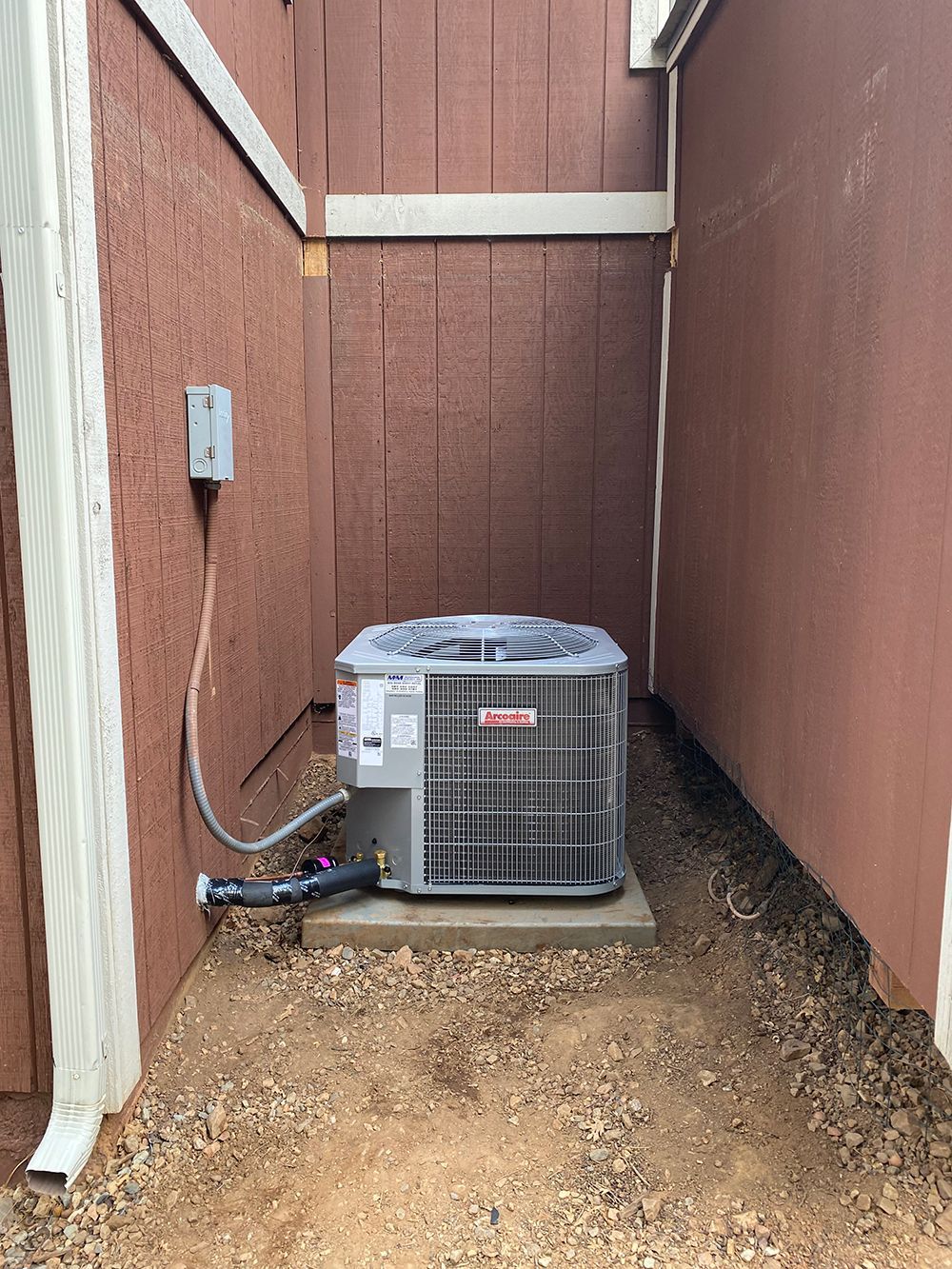 A Goodman air conditioning unit sits on a concrete pad between two exterior brown walls.