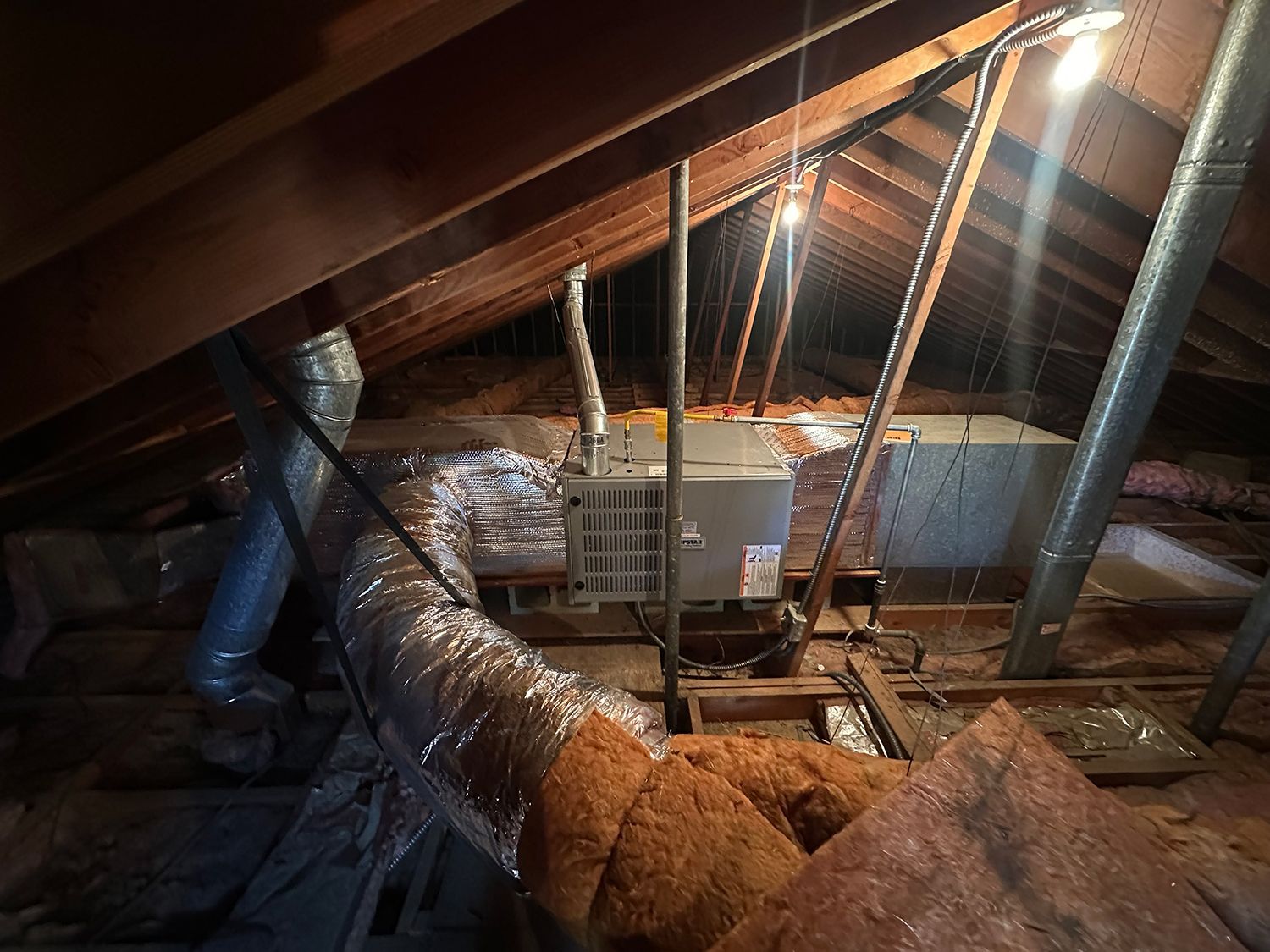 An HVAC unit and flexible metal ductwork located in a dimly lit, wooden-framed attic.