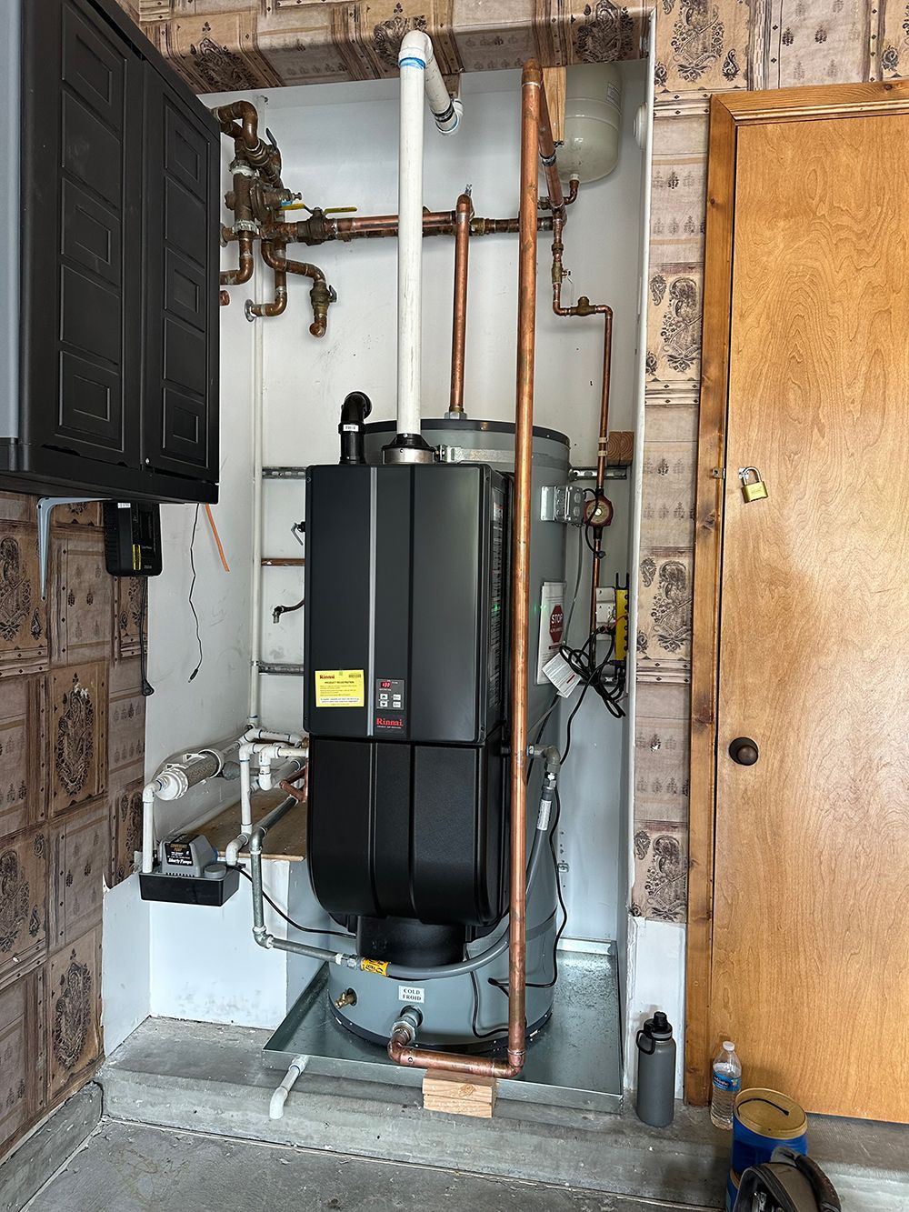 A tall, gray water heater with a black control unit, positioned in a utility closet with copper piping and a wooden door.