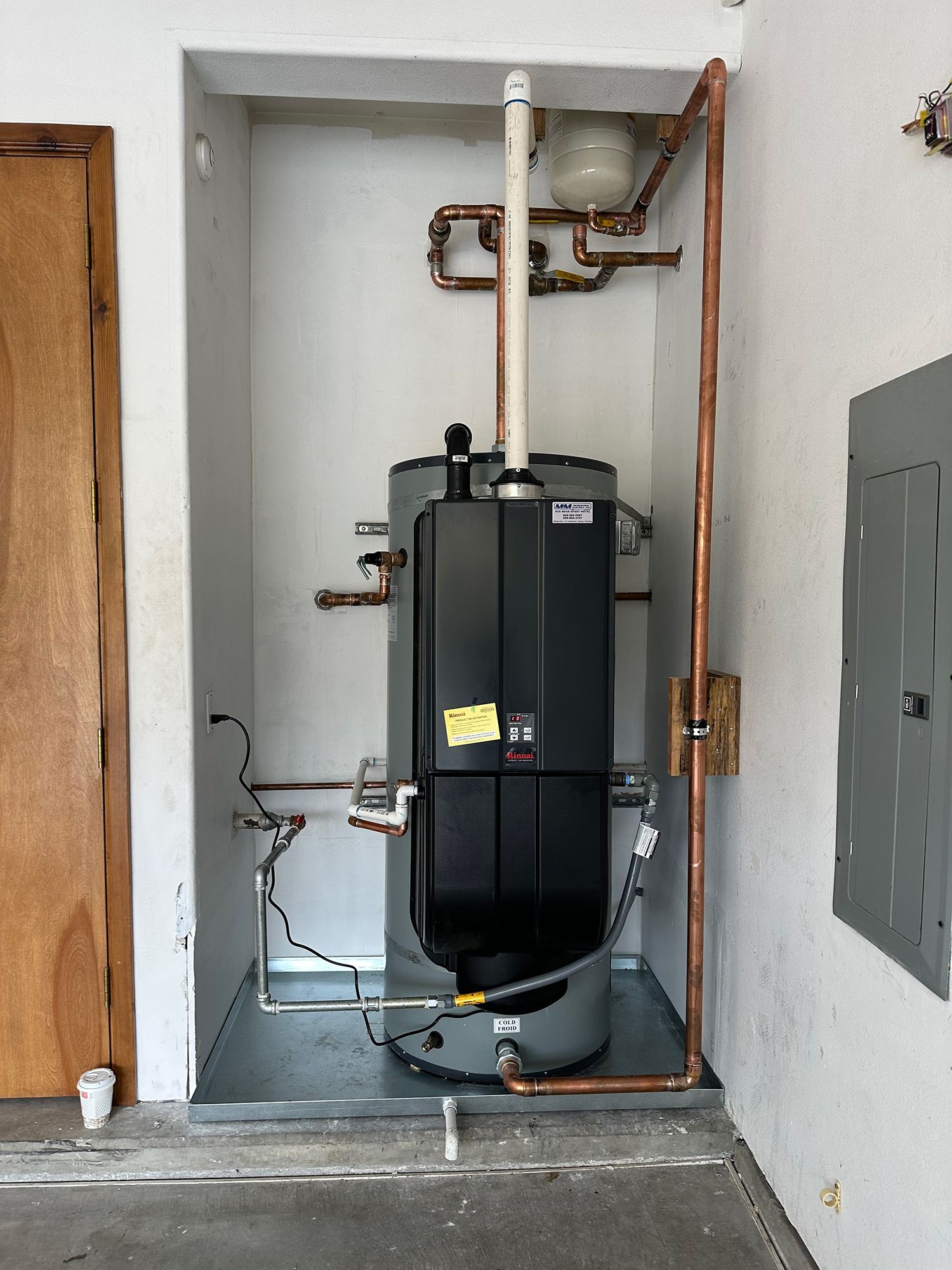 A gray water heater with black controls and copper piping sits in a recessed alcove next to an electrical panel.