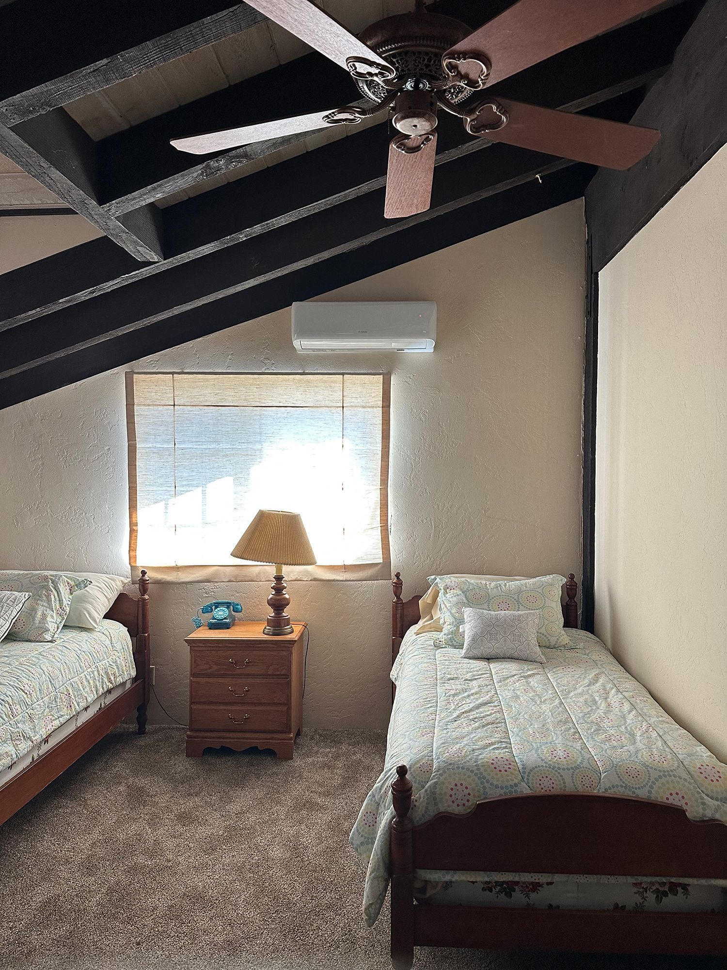 A bedroom with two twin beds, patterned quilts, a nightstand with a lamp, a ceiling fan, and a wall-mounted AC unit.