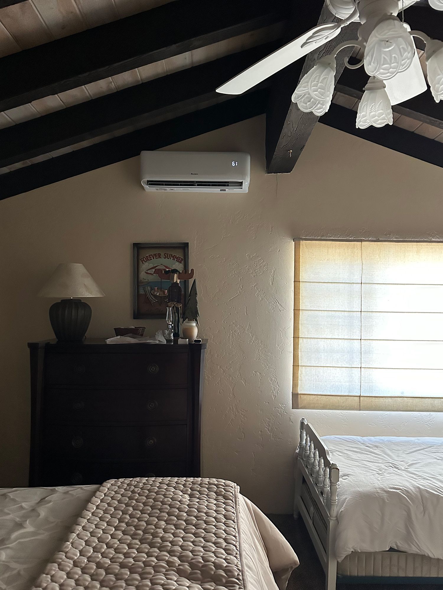 A bedroom with a slanted ceiling, ceiling fan, wall-mounted air conditioner, dark wooden dresser, and two made beds.