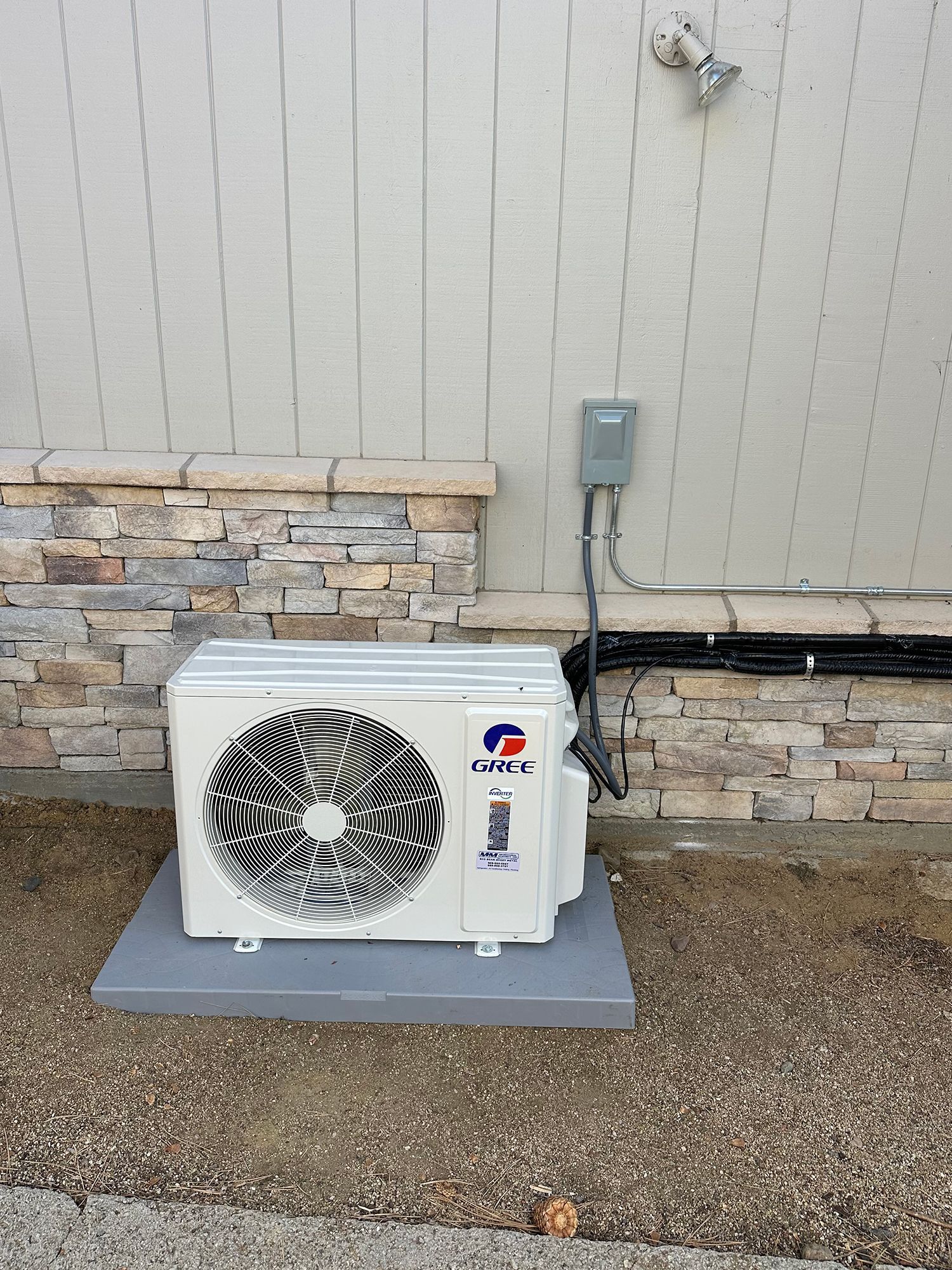 A white Gree mini-split outdoor AC unit sits on a gray concrete pad against a stone and siding wall.