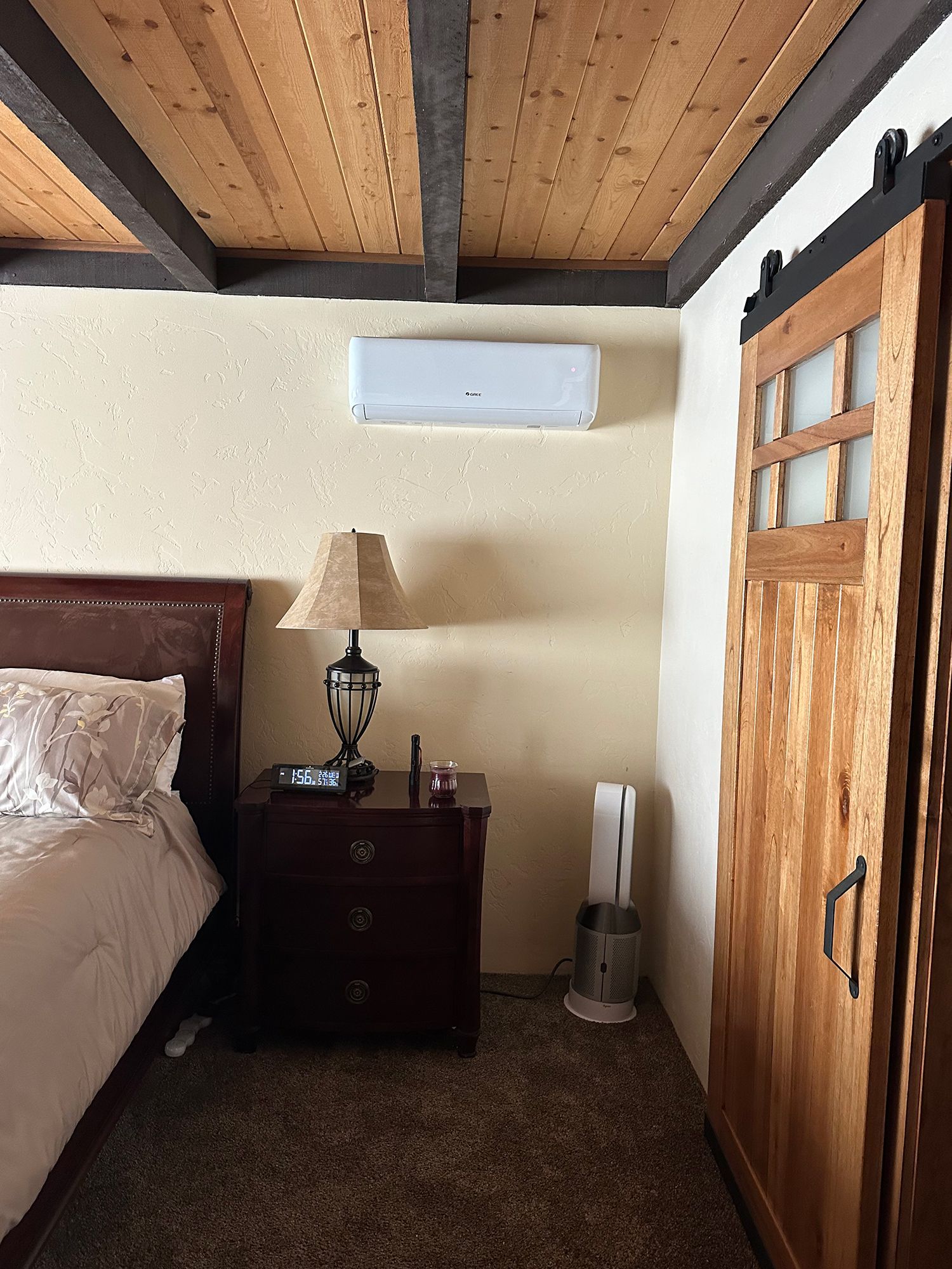 A bedroom with a wooden ceiling, a bed, a nightstand with a lamp, a wall-mounted AC unit, and a rustic sliding wooden door.
