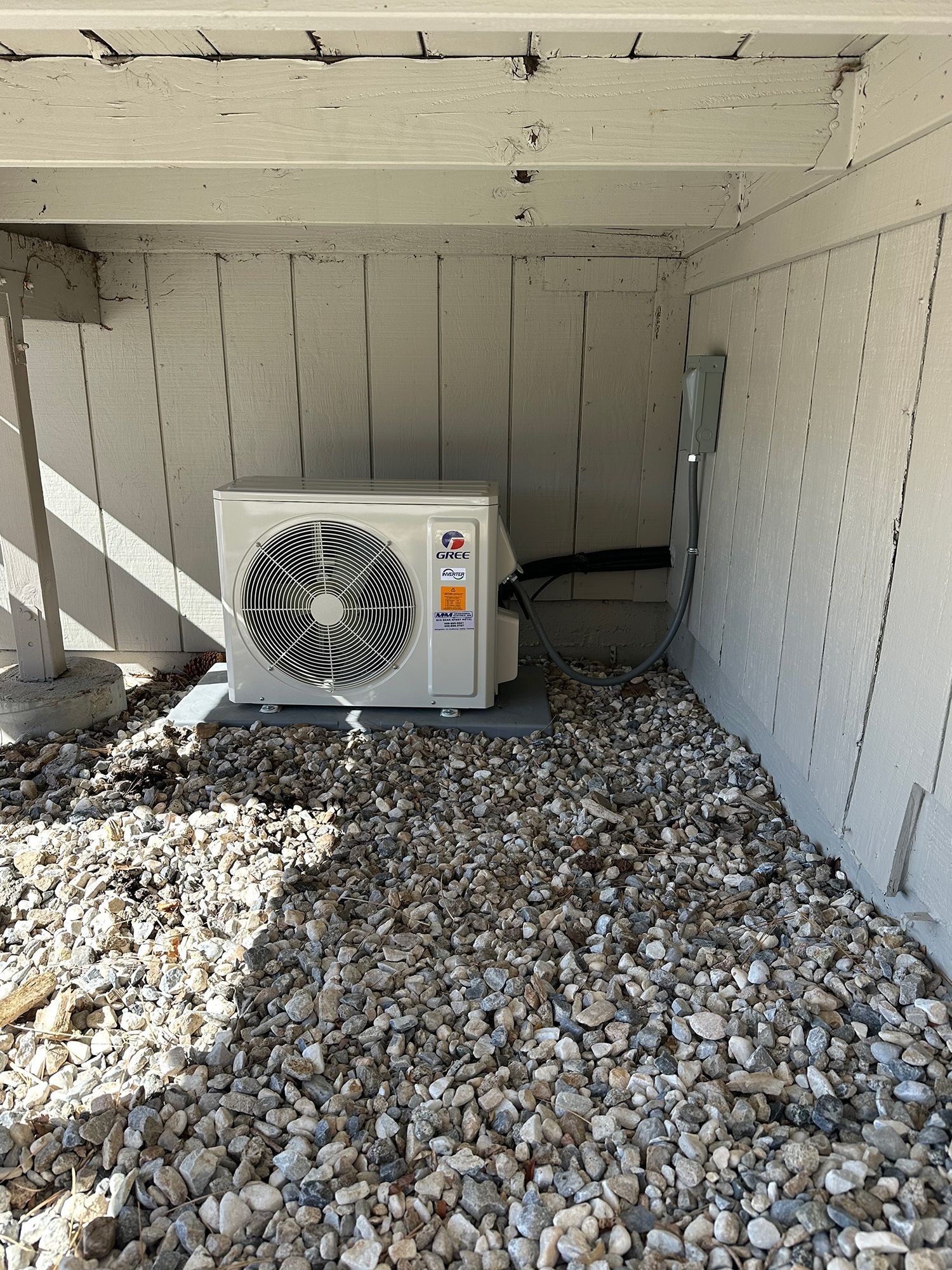 A white mini-split air conditioning unit sits on a gravel patch under a wooden deck with a nearby wall-mounted disconnect.