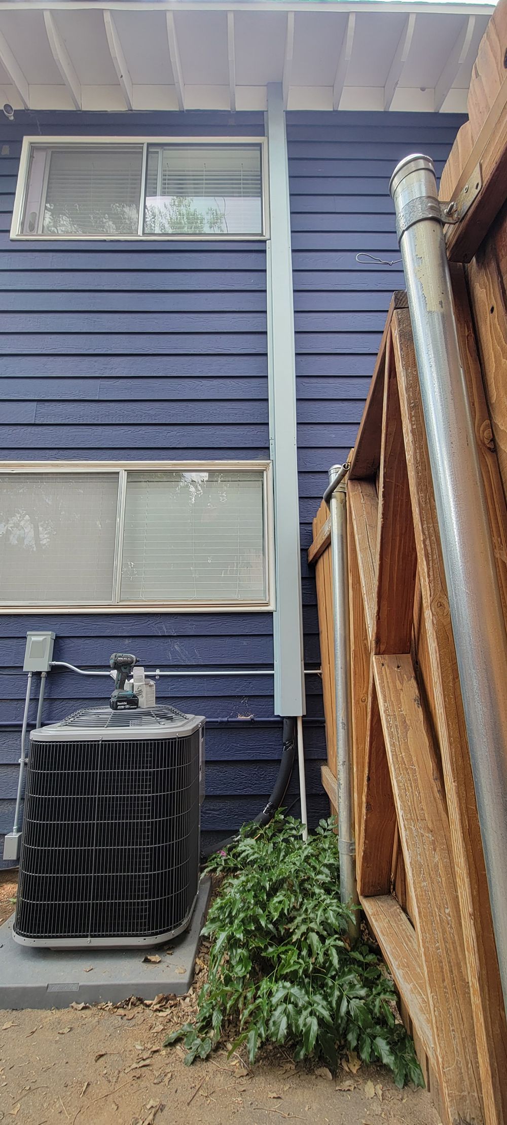A blue house exterior with a central vertical utility conduit leading to an HVAC condenser unit by a wooden fence.