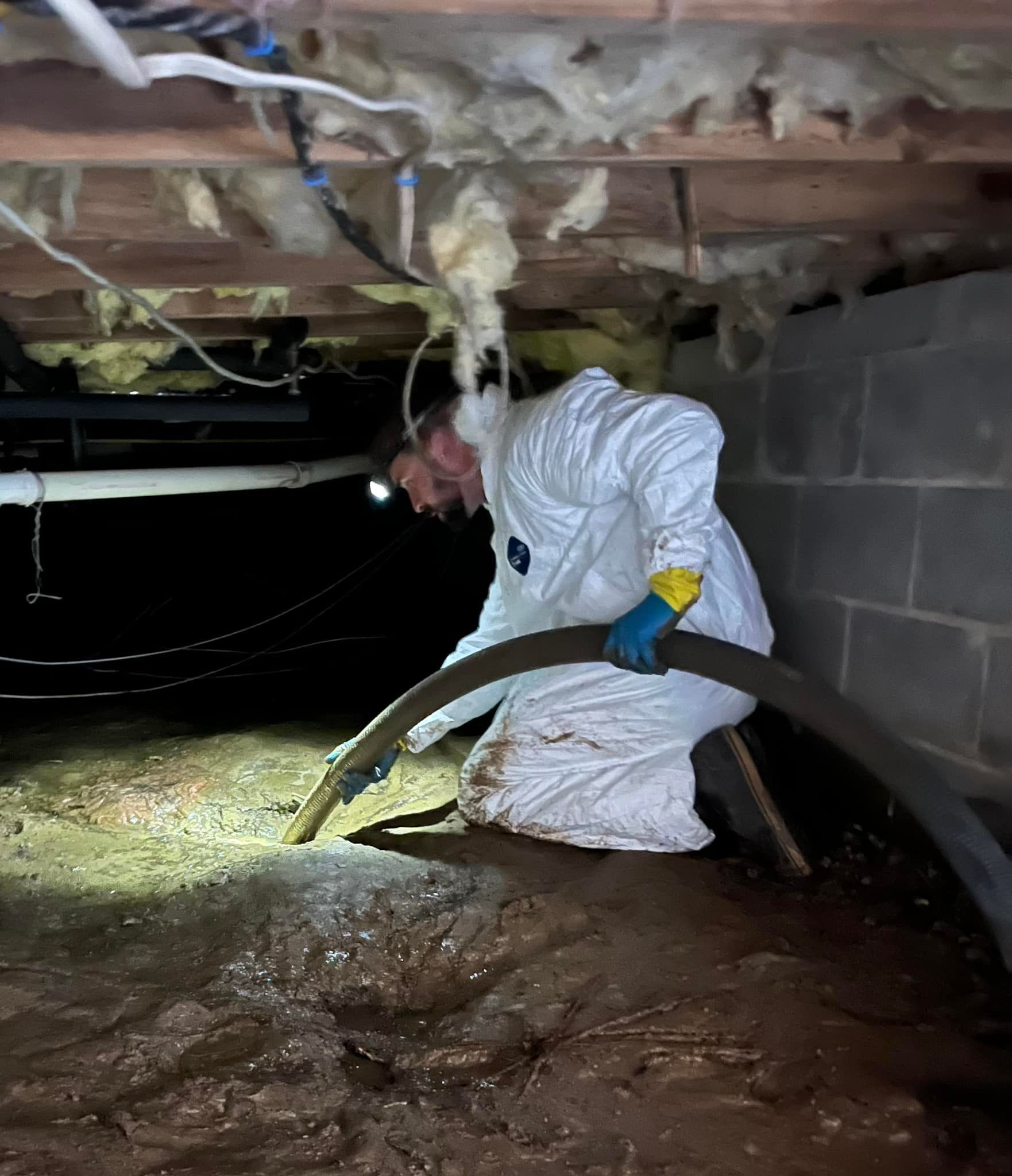 A man in a protective suit is kneeling down in a basement with a hose
