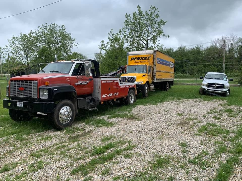 A red tow truck is towing a yellow truck in a gravel lot.