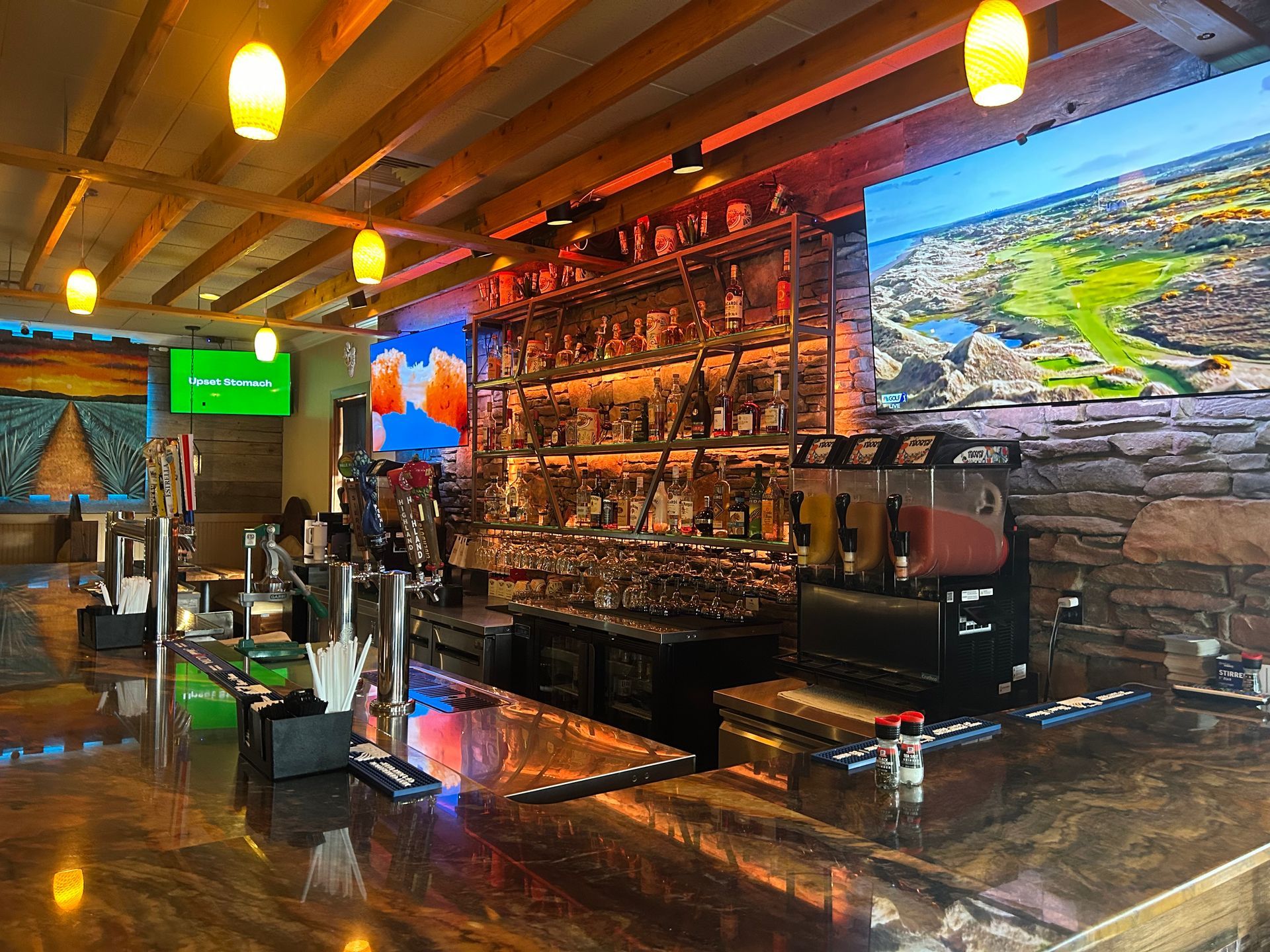Bar interior: dark wood, liquor bottles, multiple taps, TV screens, stone wall, and hanging lights.