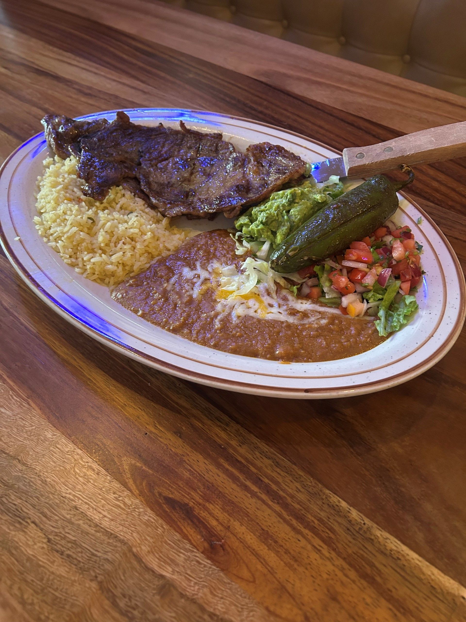 Steak dinner plate with rice, beans, guacamole, salsa, and a pickle on a wood table.