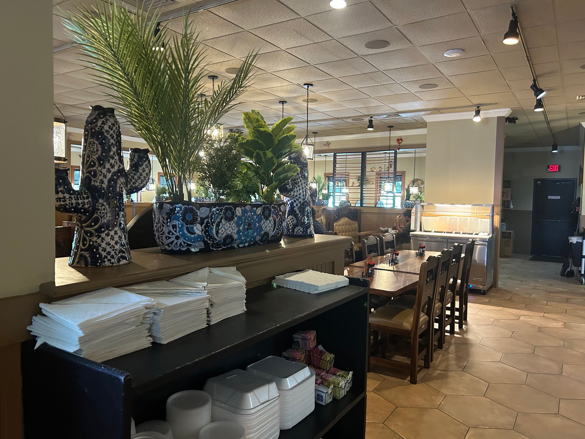 Restaurant interior with tables, chairs, decorative cactus, plants, and a shelf with napkins and containers.
