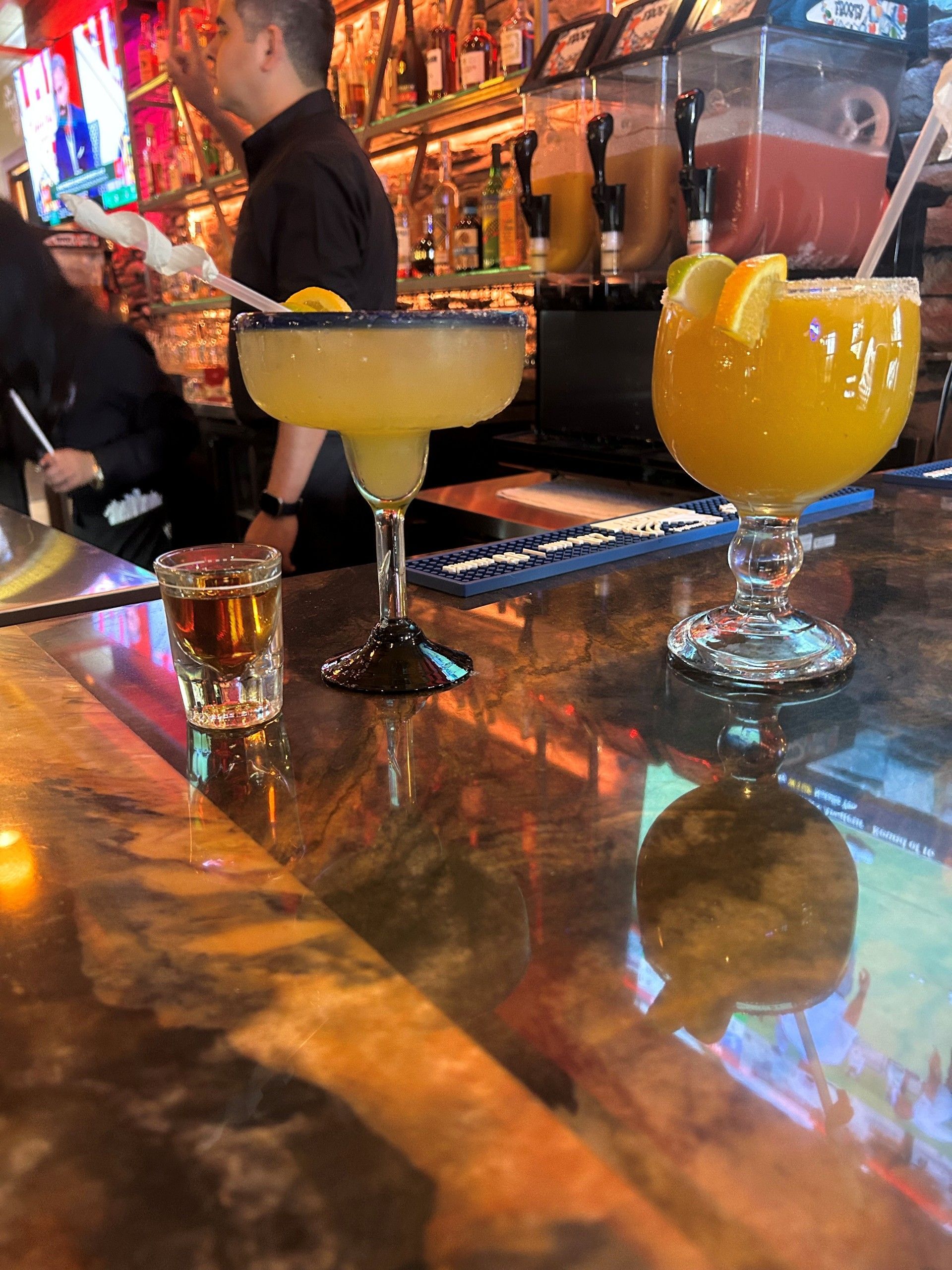 Margarita and orange cocktail on a bar, shot of liquor, person behind the bar, bar setting.