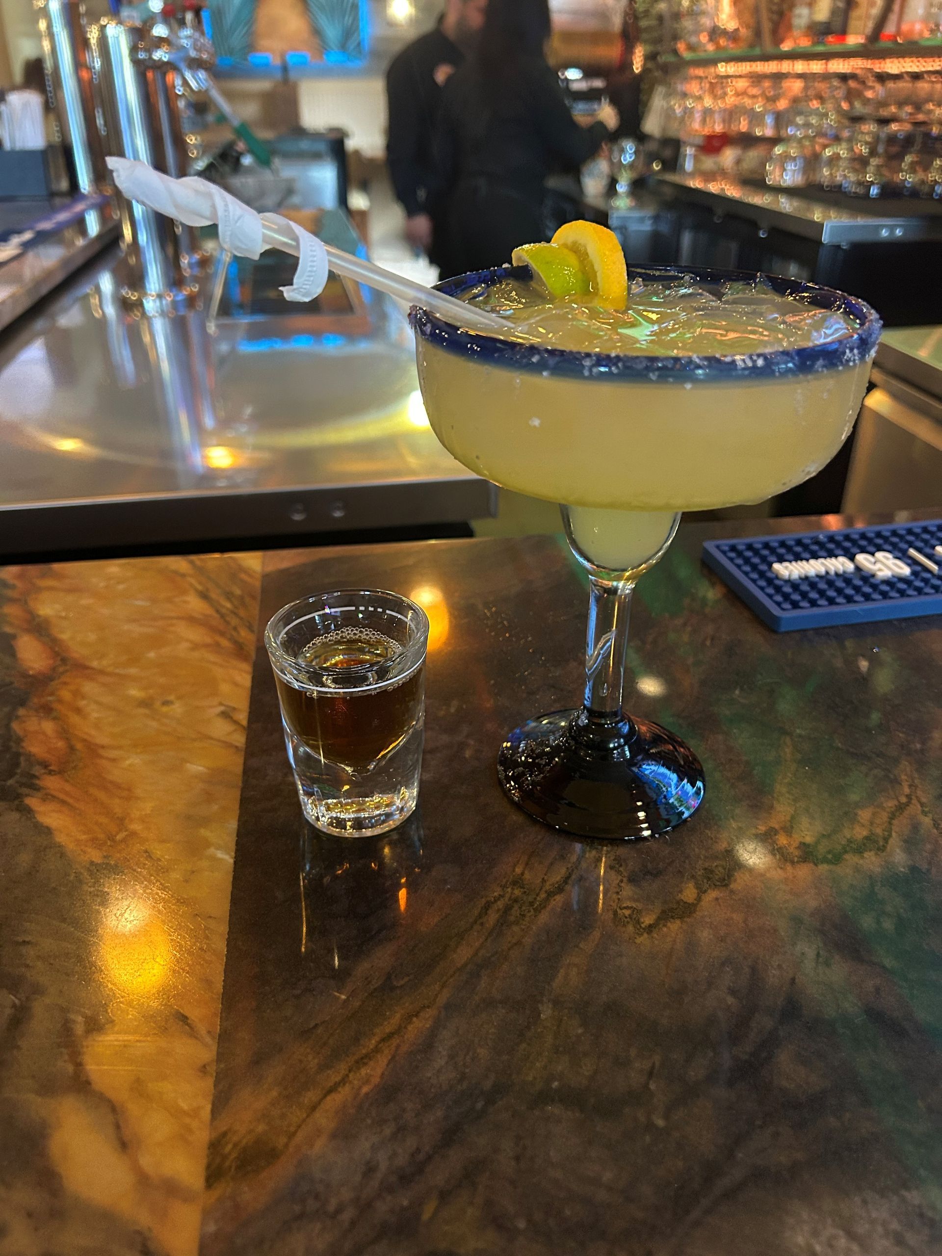 Margarita and shot of liquor on a bar. Margarita with lime garnish, blue rimmed glass. Shot of liquor beside it.