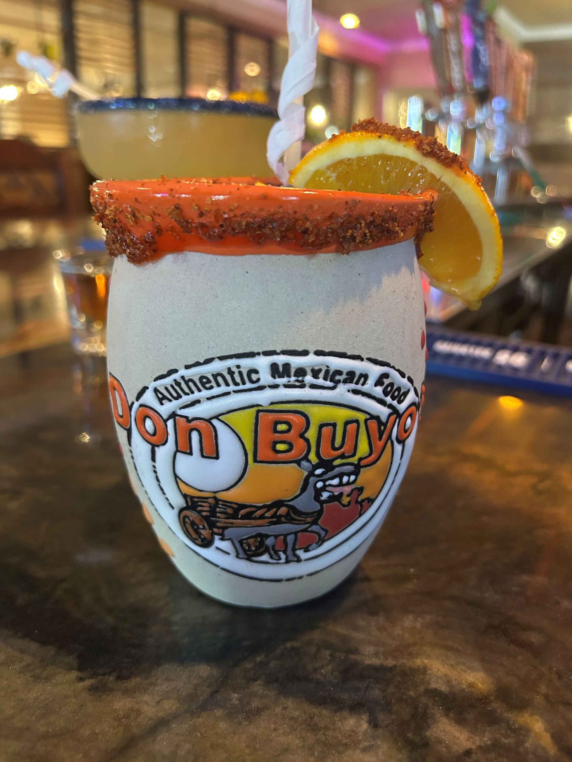 A colorful cocktail in a branded, rim-salted mug, garnished with orange slice. Bar setting.