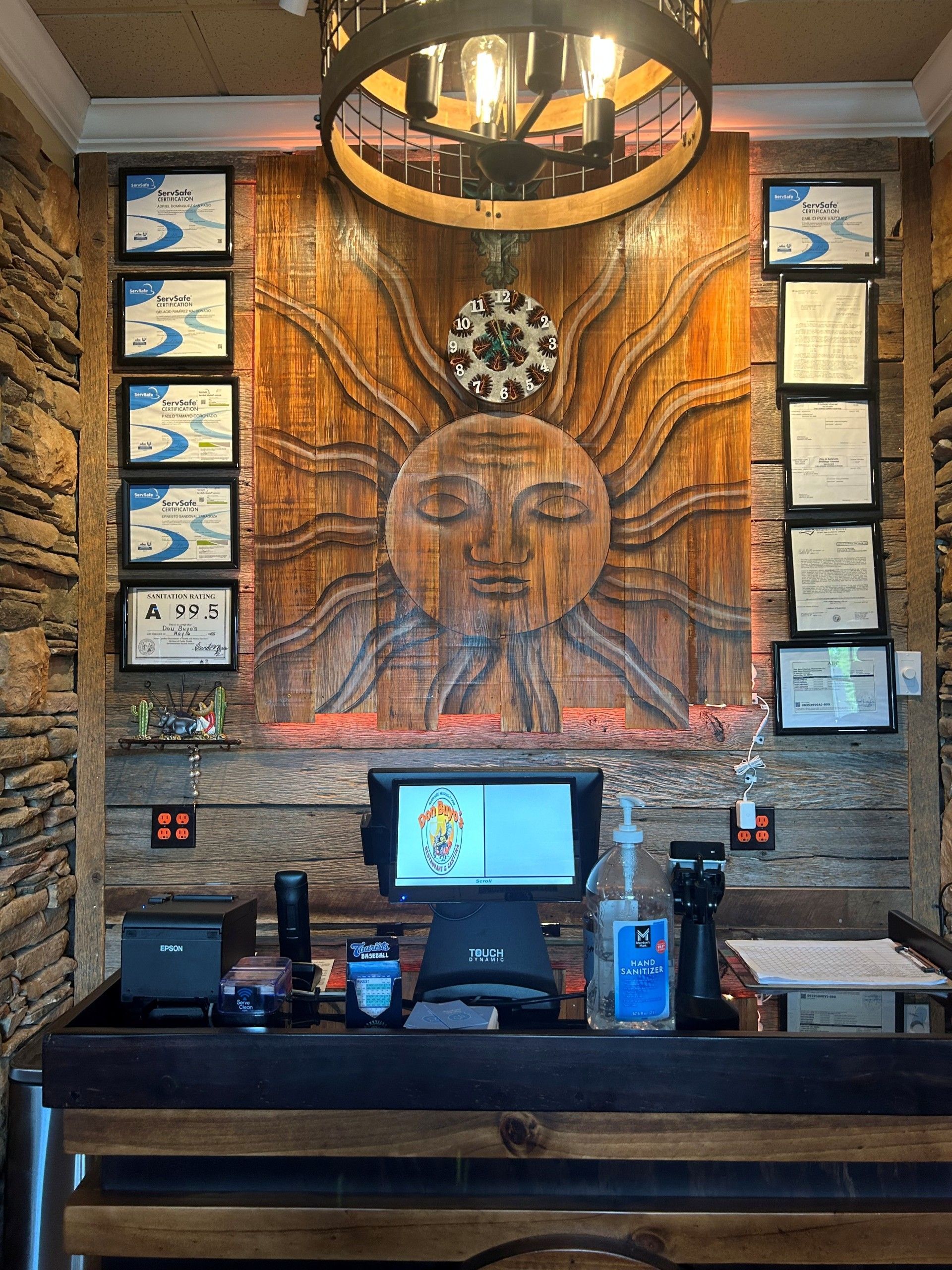 Cashier station with sun mural backdrop, surrounded by stone, framed certifications, and a chandelier.