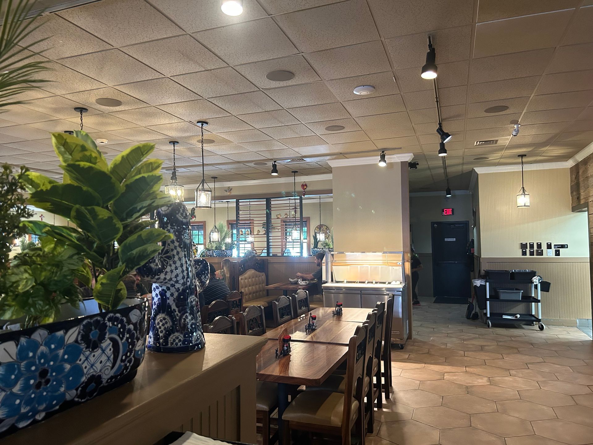 Restaurant interior with wooden tables and chairs, plants, and a black door at the end of the room.