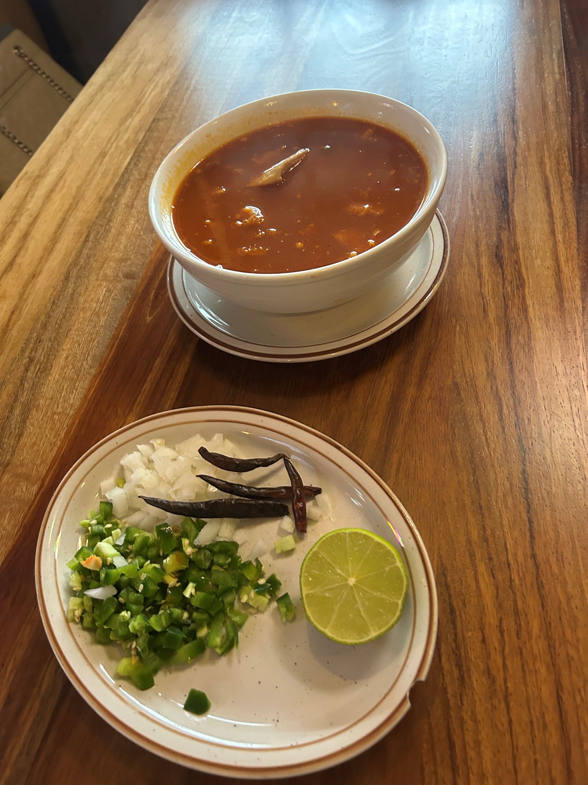 Bowl of red soup with a side plate of chopped jalapeños, onions, dried chilies, and a lime half on a wooden table.