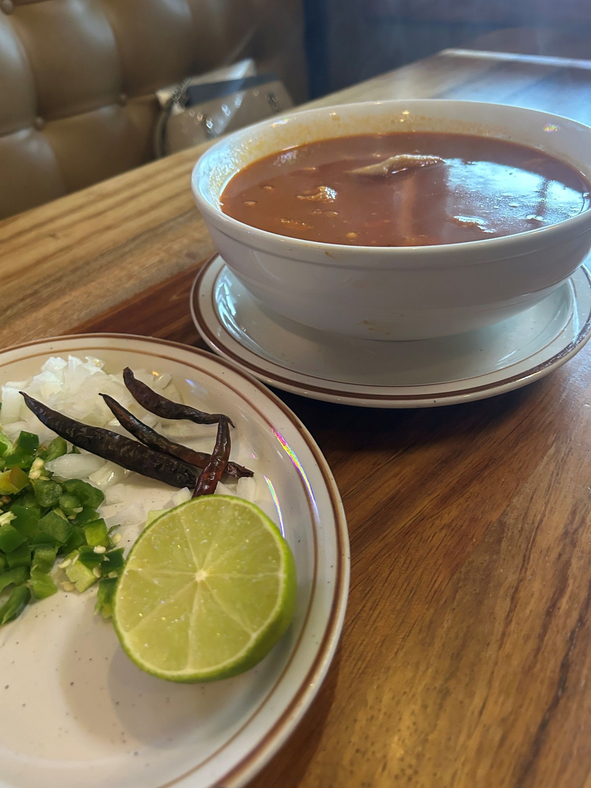 Soup bowl and plate with chopped onions, jalapeños, lime, and dried peppers on a wooden table.