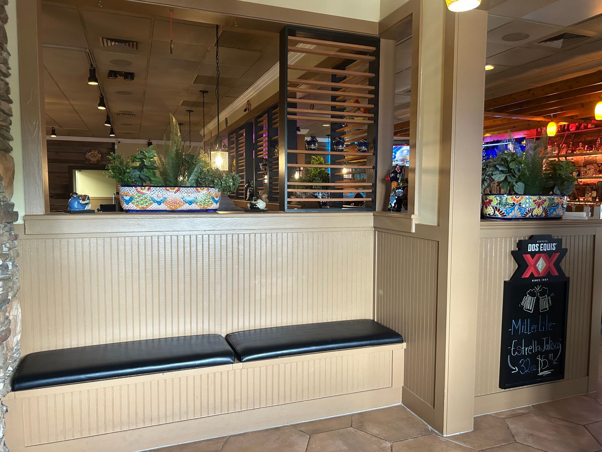 Booth seating in a restaurant. Beige walls, black cushioned bench, planters with greenery.