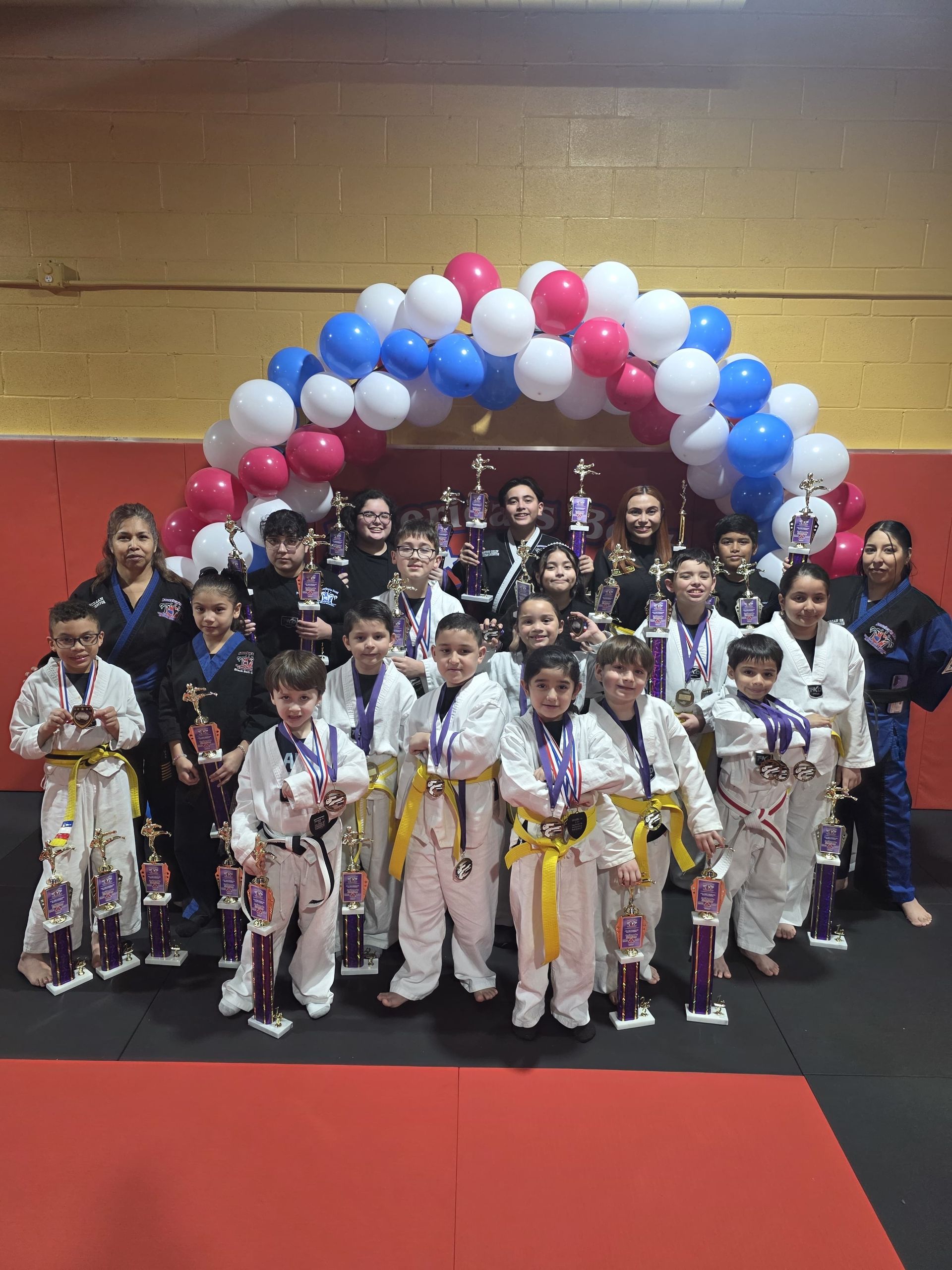 Group of martial arts students holding trophies and medals under a balloon arch.