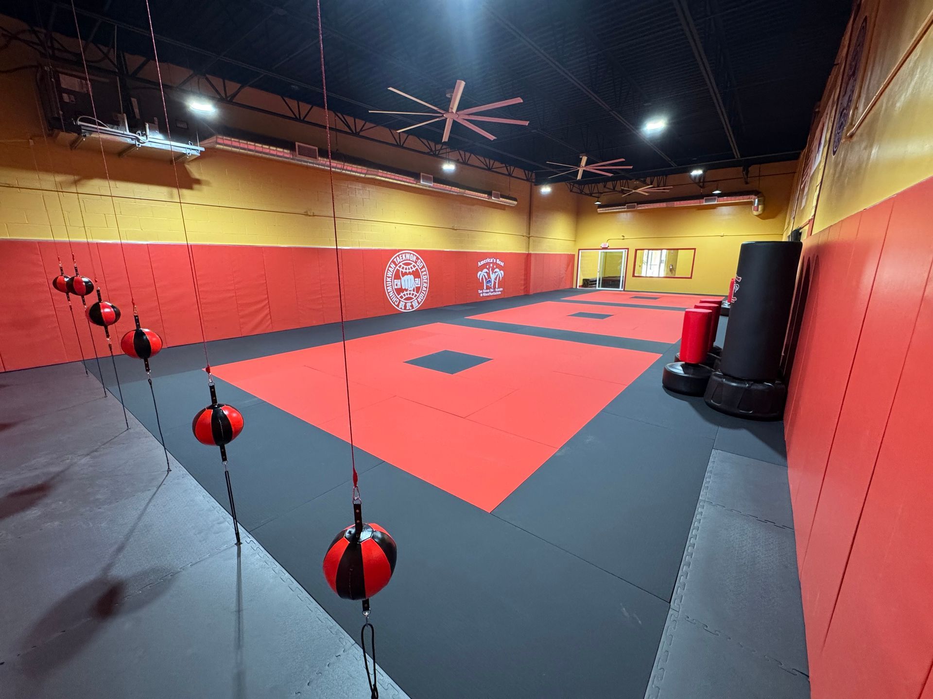 Martial arts training room with red and black mats, punching bags, and hanging speed balls.