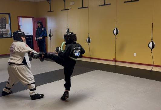 Two people sparring in a martial arts studio. One kicks, the other blocks. Yellow wall, black bags hanging.