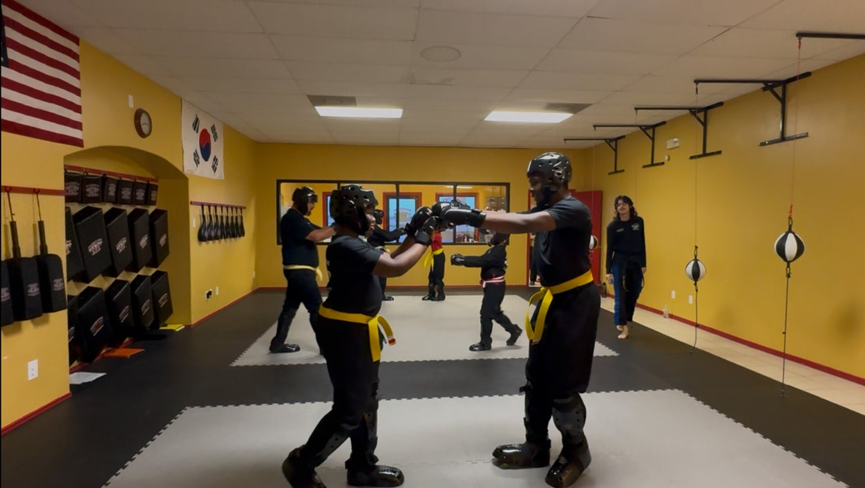 Martial arts class: Students sparring, wearing protective gear, in a dojo with yellow walls, flags, and training equipment.
