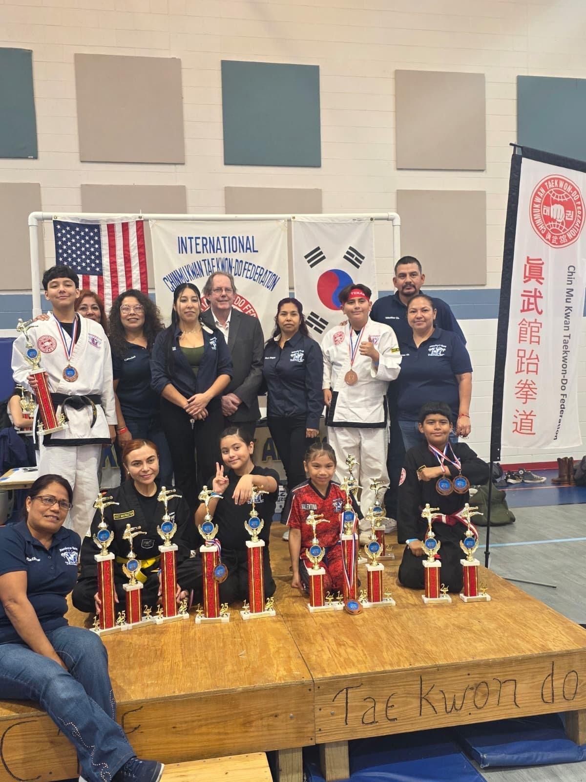 A group of Taekwondo practitioners in uniforms and casual clothes posing with trophies on a wooden stage indoors.