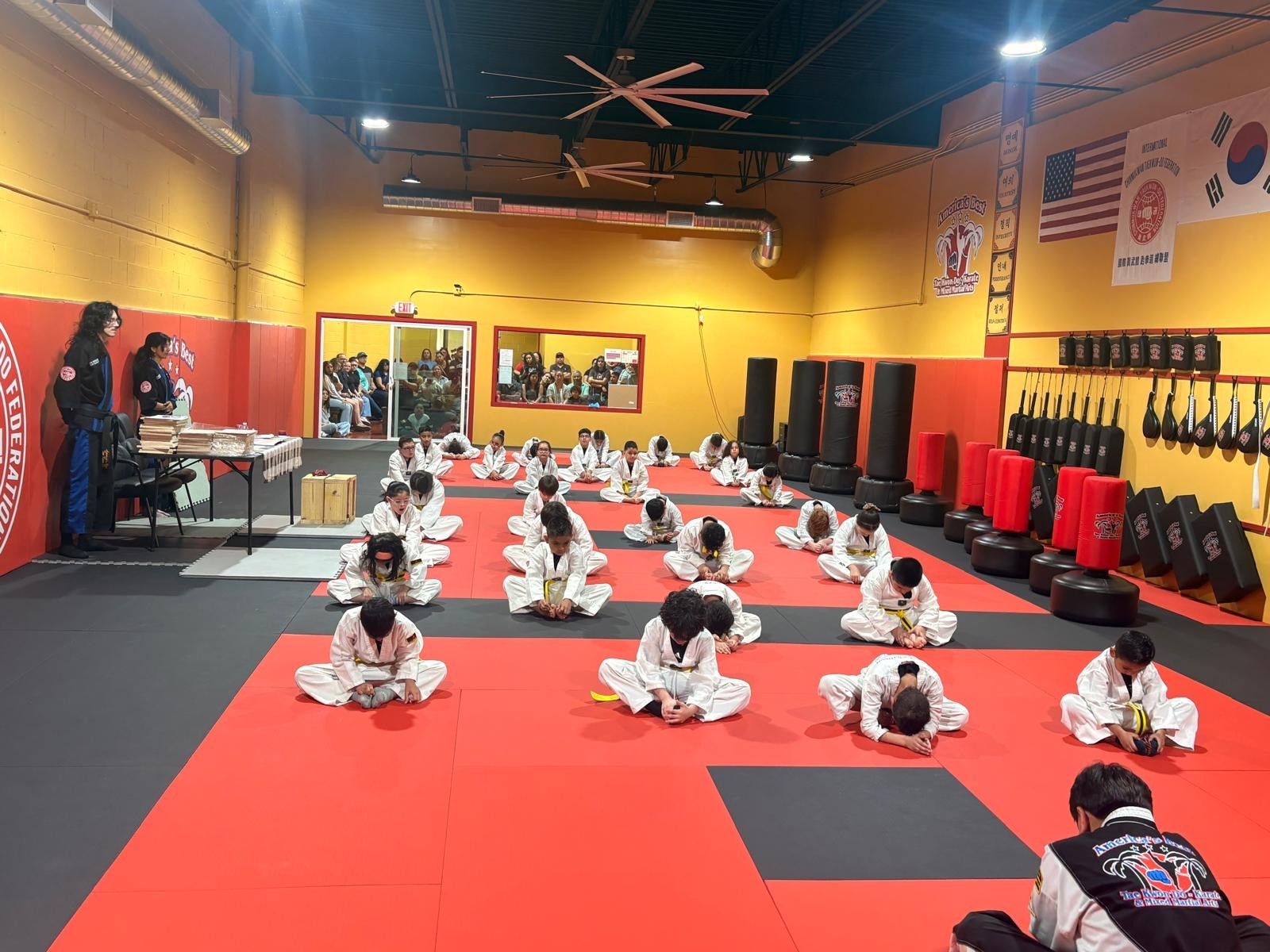 Students in white uniforms sit on a red mat in a martial arts studio, stretching during a class.