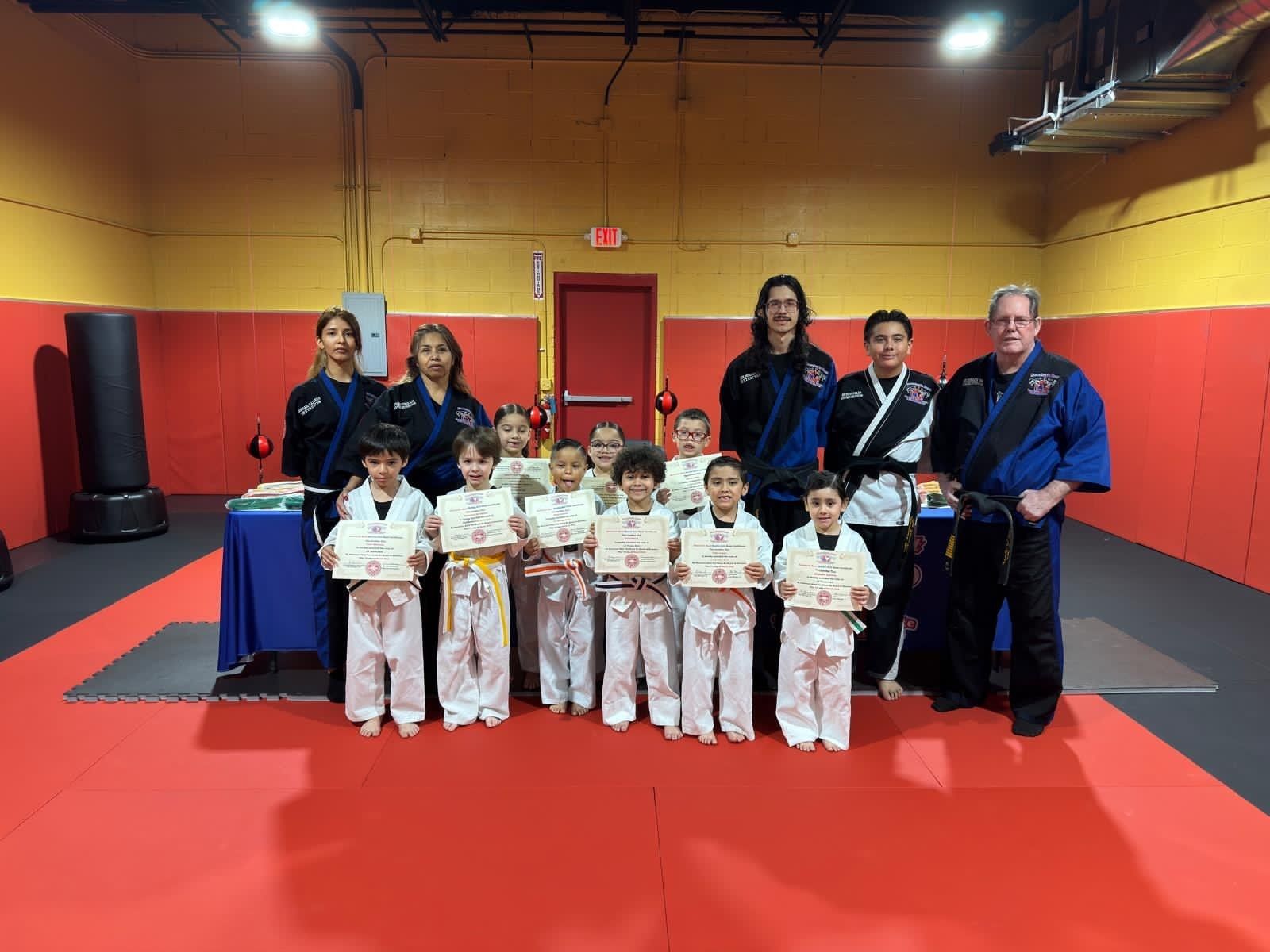 A group of children and instructors in martial arts uniforms stand indoors, holding certificates after a class.