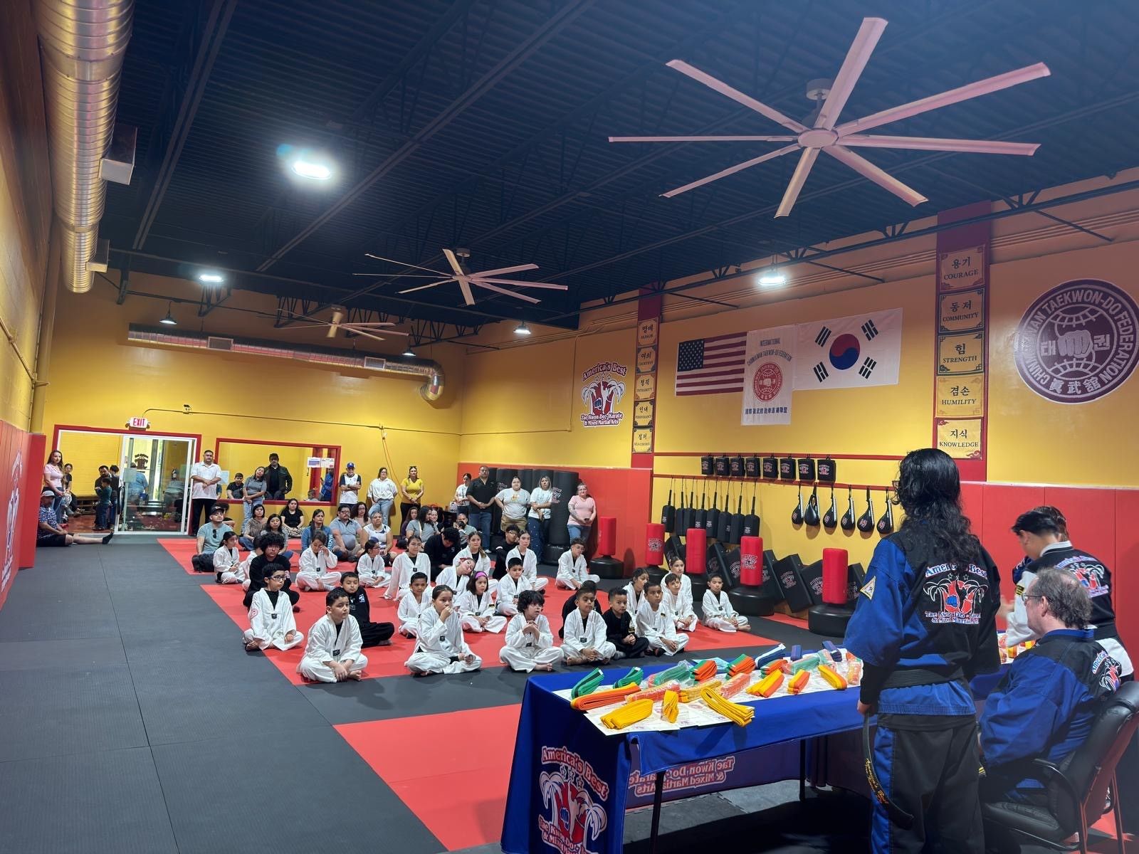A Taekwondo class sits on floor mats in a studio with American and South Korean flags, facing an instructor at a table.