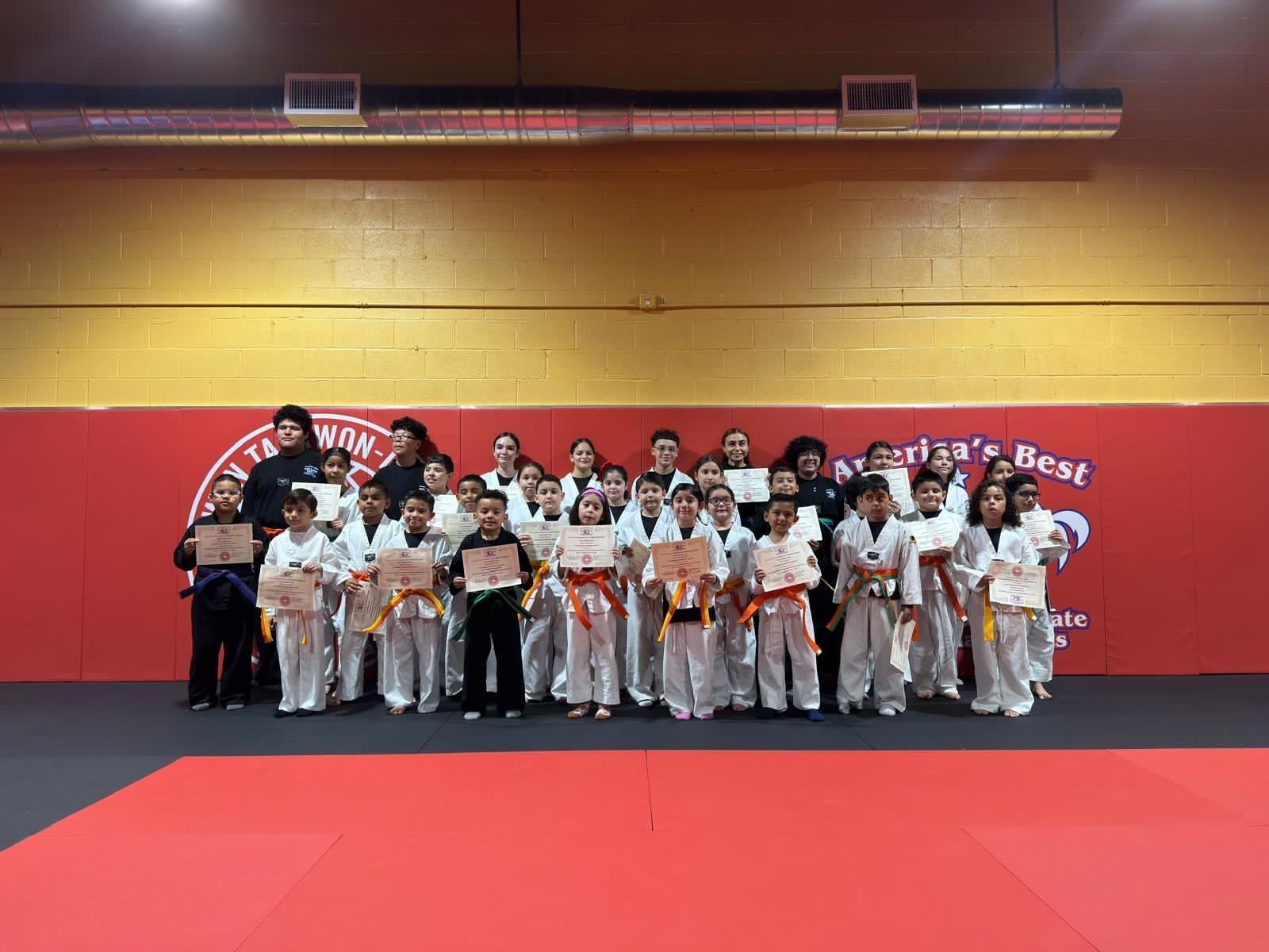 A group of students in martial arts uniforms stand in front of a red wall holding certificates at a taekwondo studio.