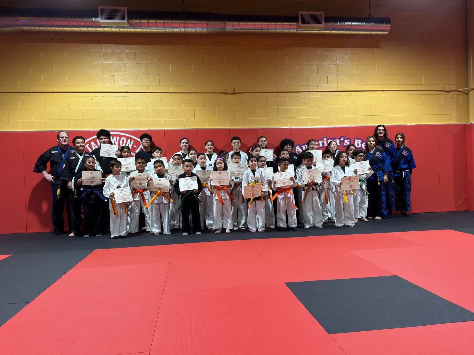A large group of martial arts students in white uniforms holding certificates, lined up in a gym with red mats.