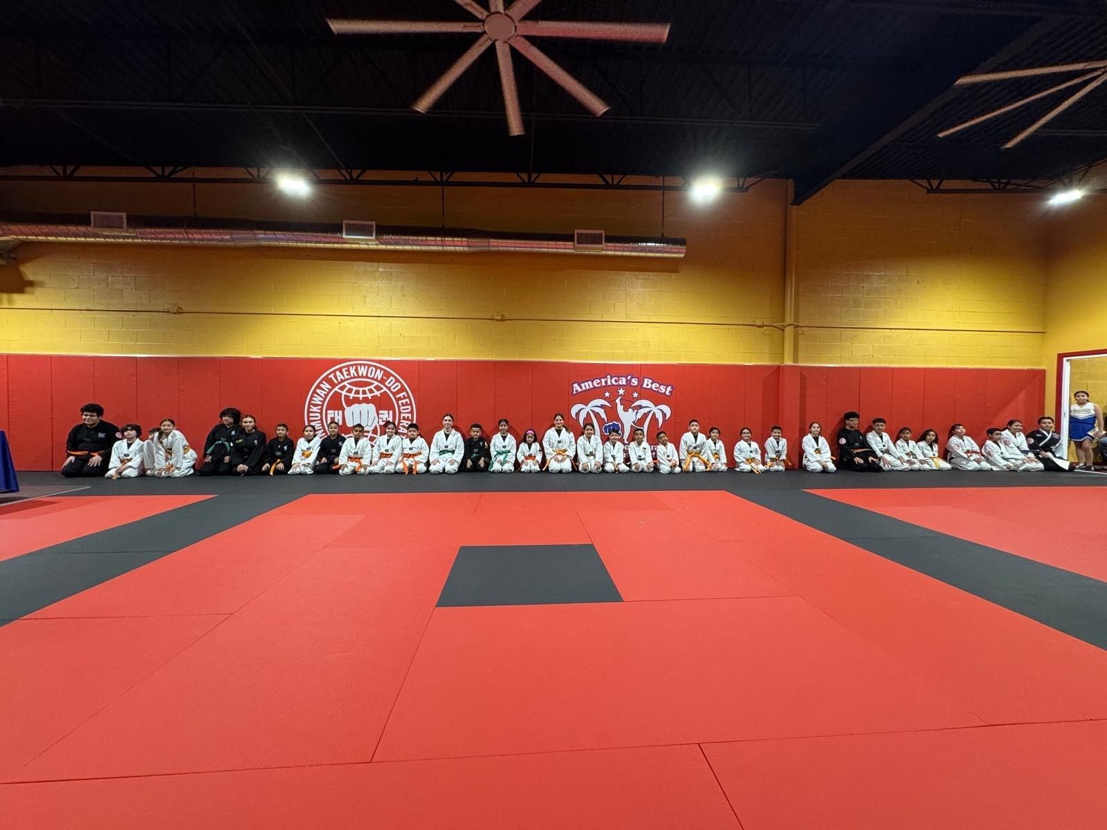 A group of people in martial arts uniforms sit in a line on a large red and black mat in a training studio.