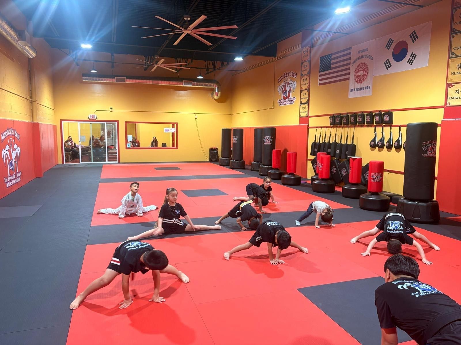 Students in black and white uniforms perform stretching exercises on red and gray mats in a martial arts studio.