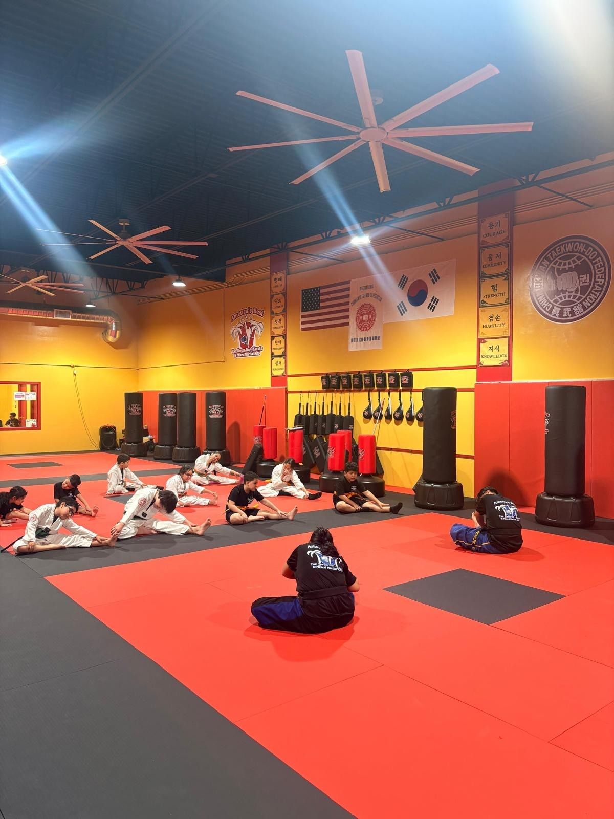 A group of people wearing martial arts uniforms stretches on red and gray gym mats in a training studio.