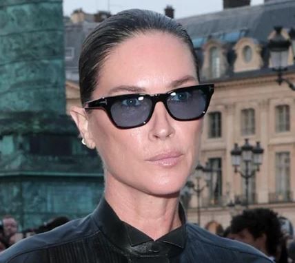 Person in black sunglasses and dark jacket at an outdoor event, with historic buildings in the background