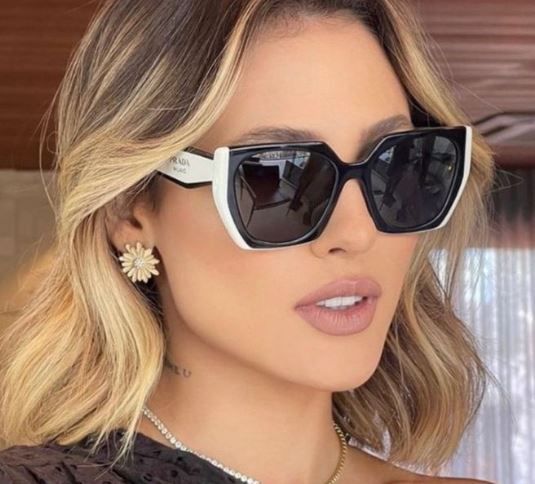 Portrait of a woman wearing black sunglasses and a dark top, with blonde wavy hair and flower earrings