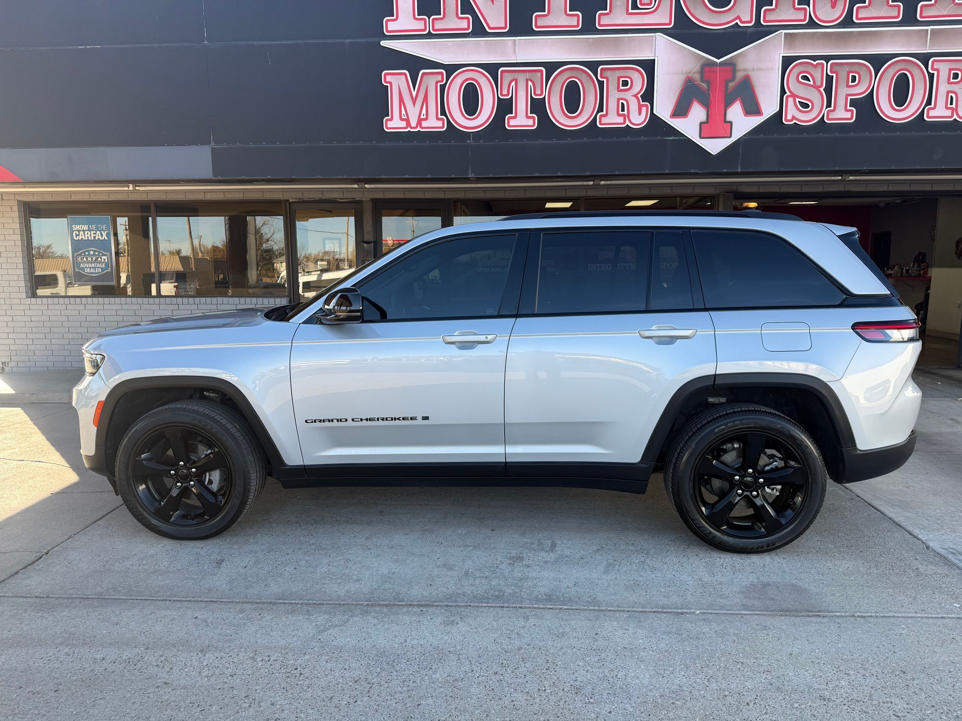 Silver Jeep SUV parked in front of a motor sports shop, black wheels, tinted windows.