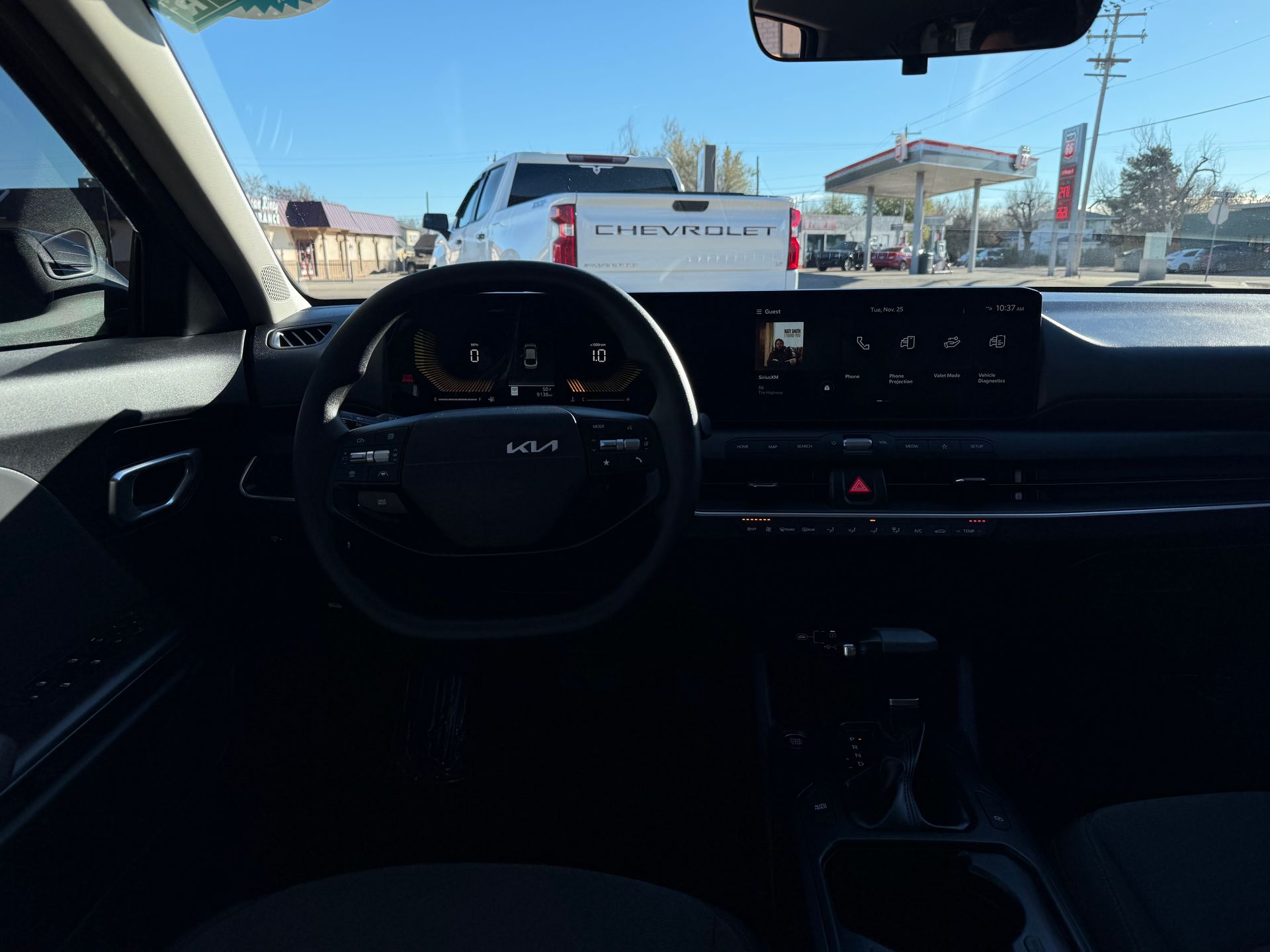 Interior view of a Kia vehicle dashboard, steering wheel visible. The car is facing a street.