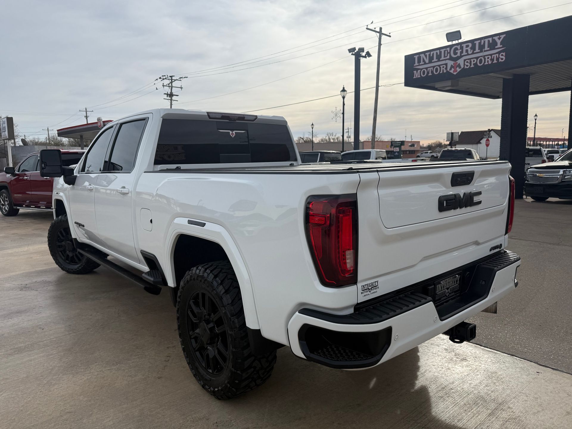White GMC truck, black wheels, parked outside a dealership, sunny day.